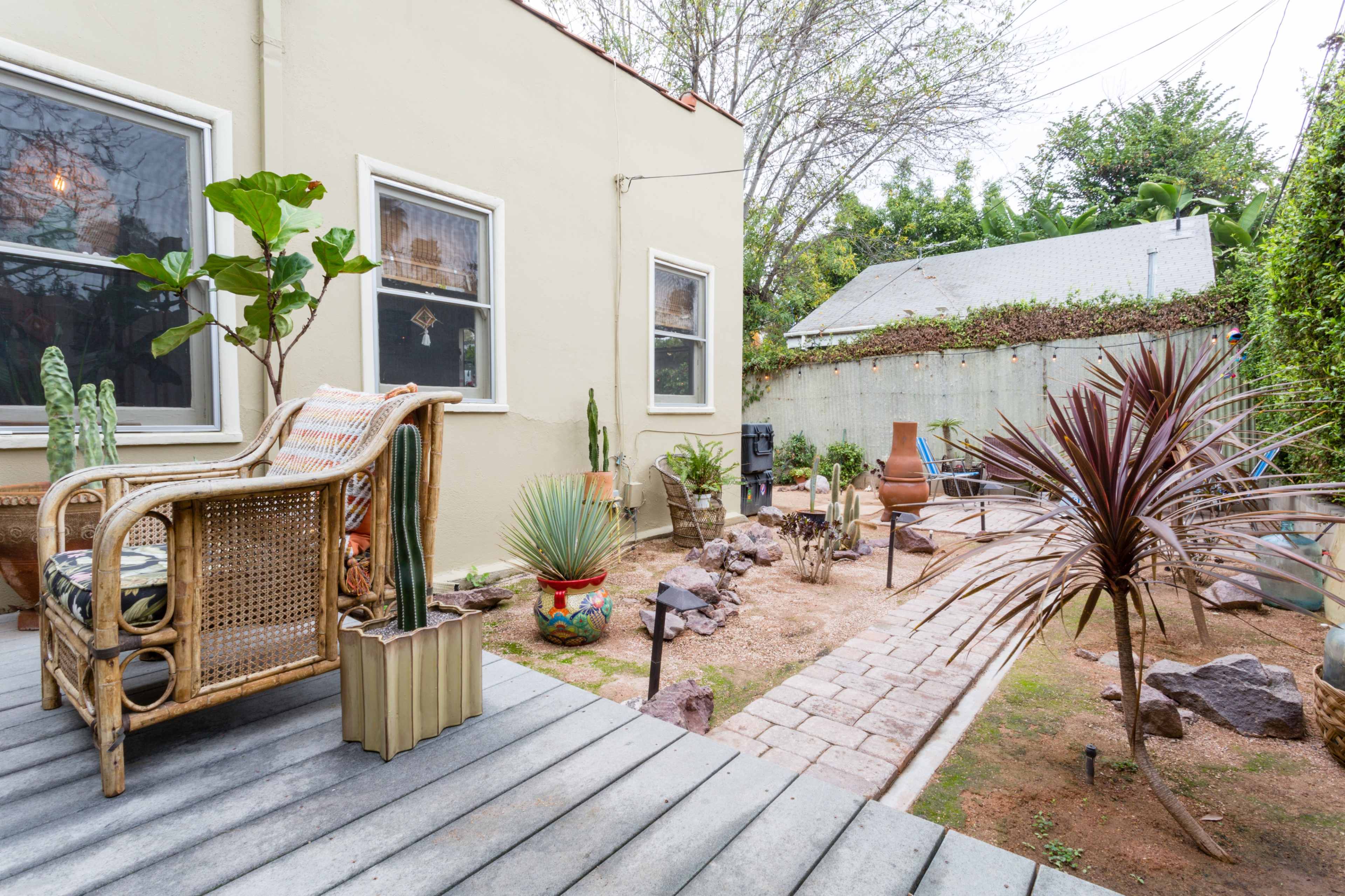A small, landscaped backyard features a seating area with a wicker chair, various plants, and a path made of stone tiles.