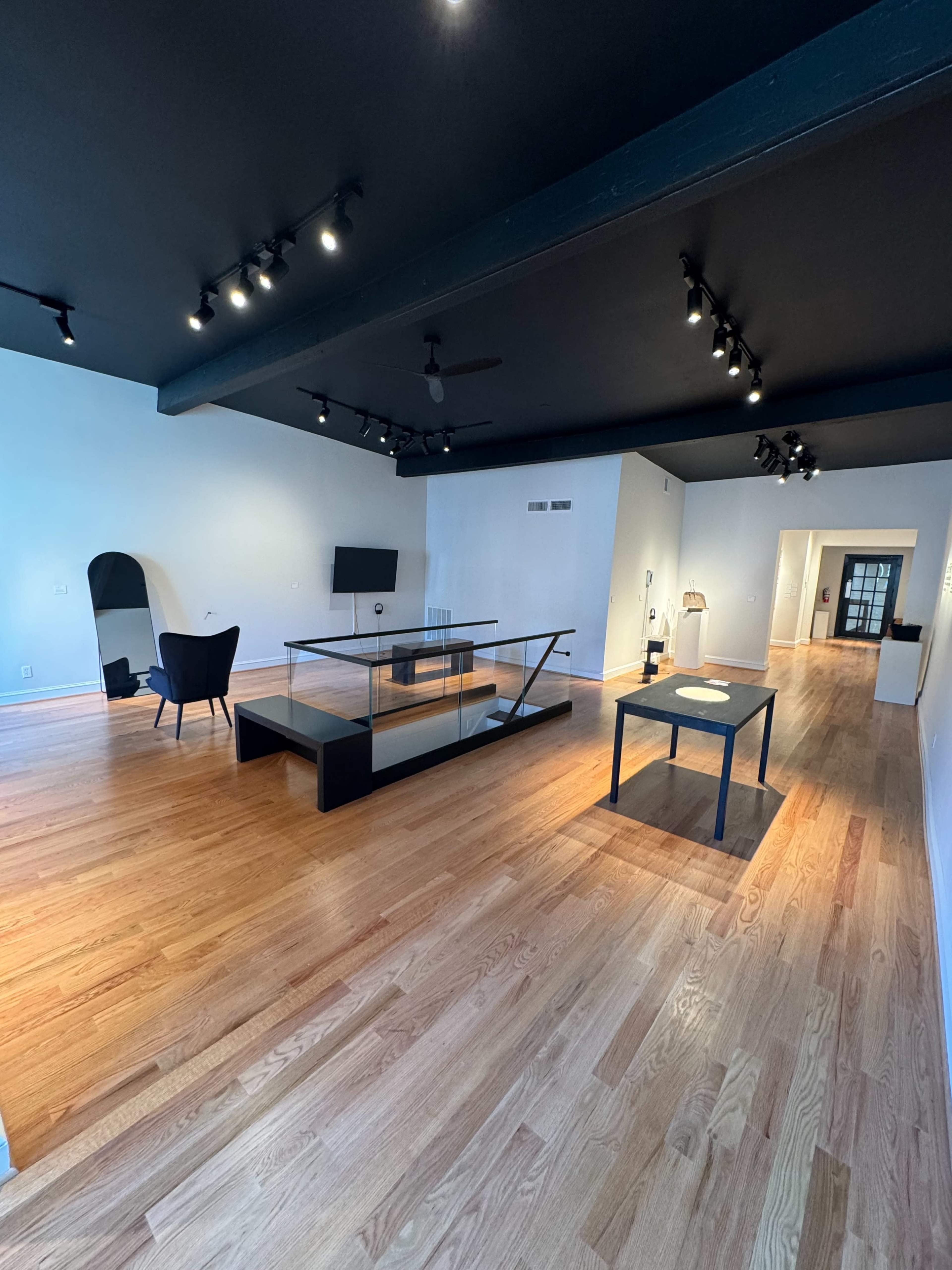 A spacious room with wooden flooring, featuring a black ceiling, minimalist furniture, and a large glass table in the center.