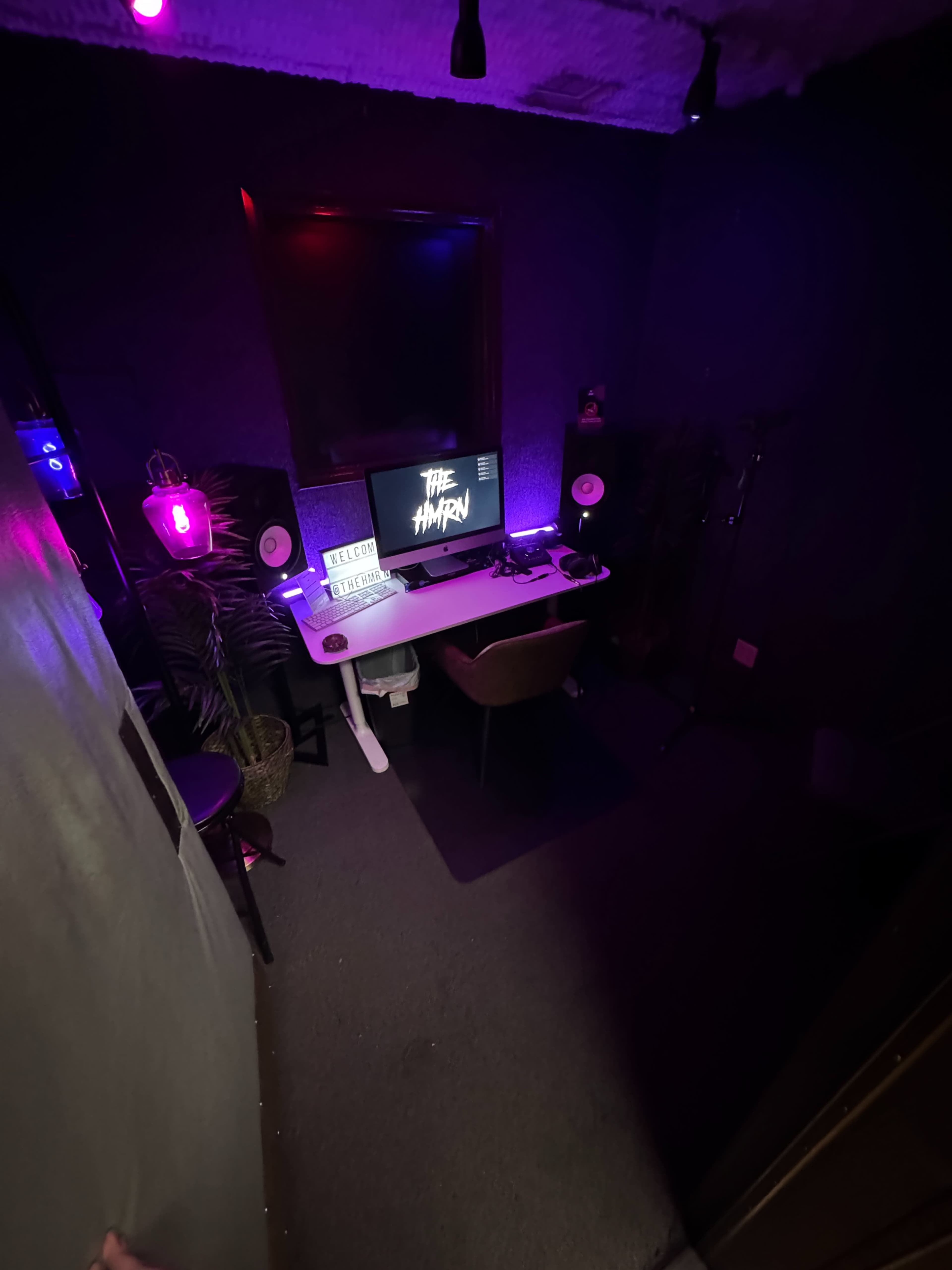 The image shows a small, dimly lit audio or recording studio with a desk, computer, and sound equipment.