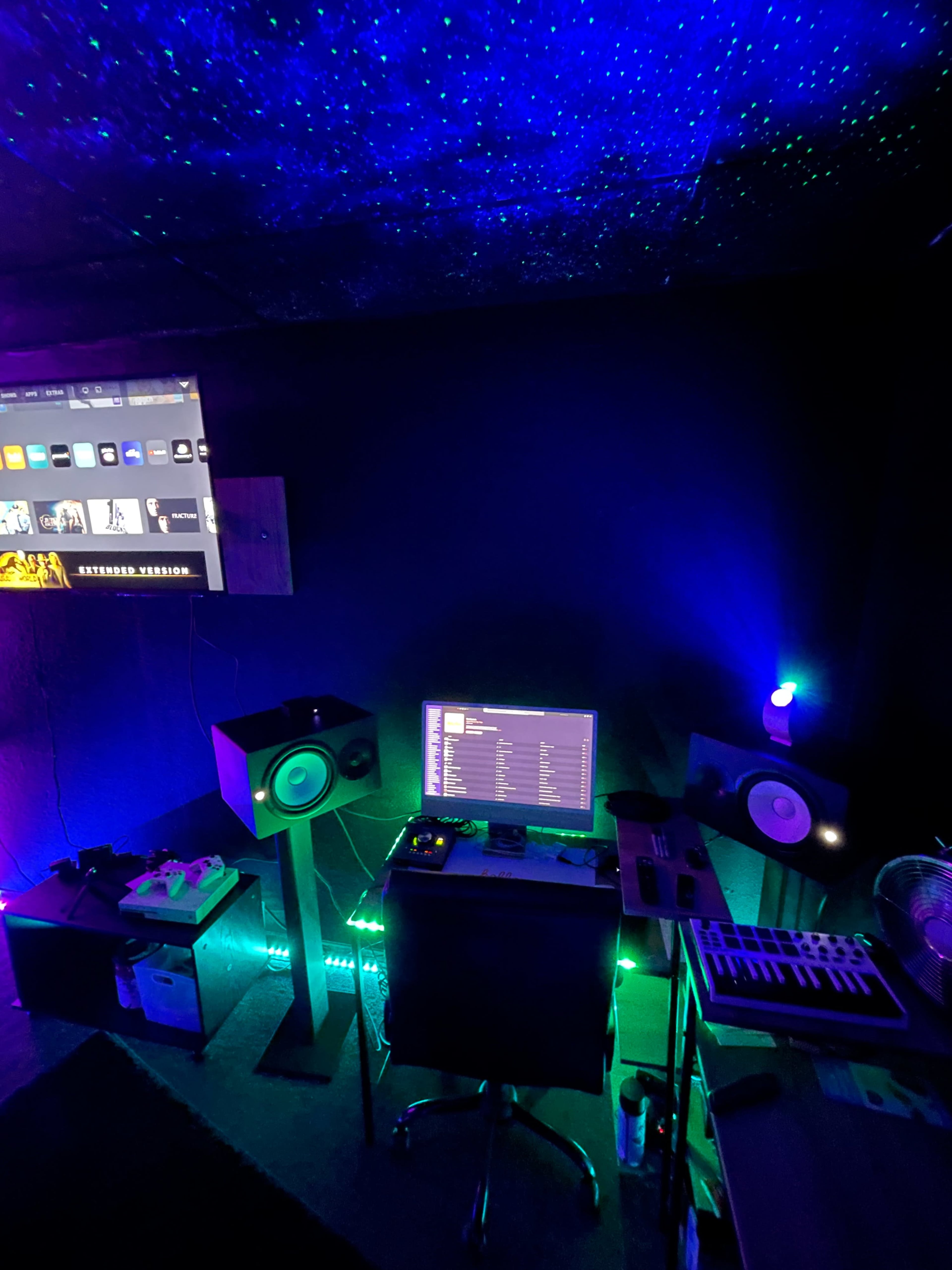 A music production studio is illuminated with colorful lights, featuring a computer, speakers, and a MIDI keyboard on a desk.