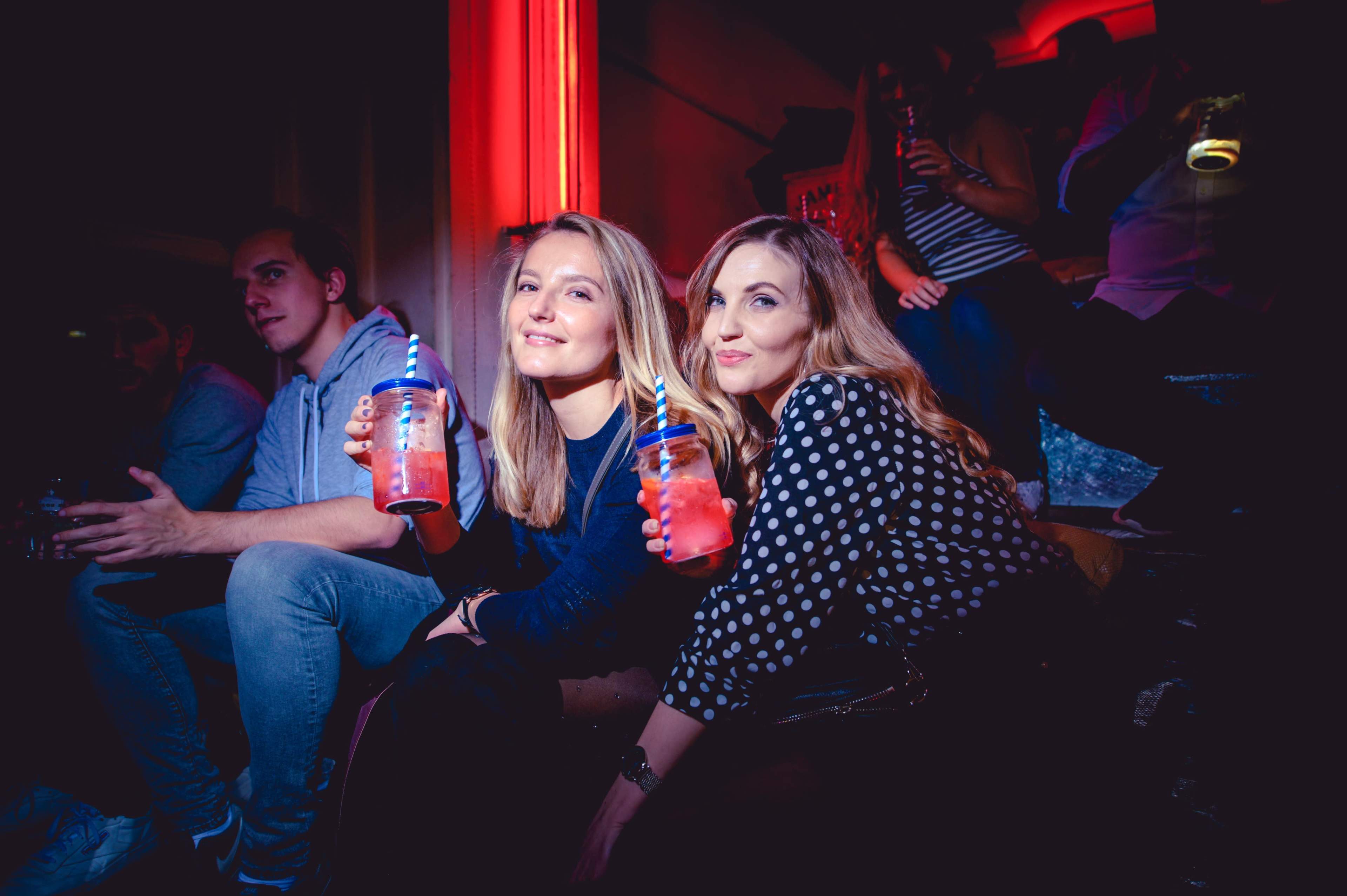 Two women sit together holding drinks in a vibrant setting with subdued lighting, while a group of people is visible in the background.
