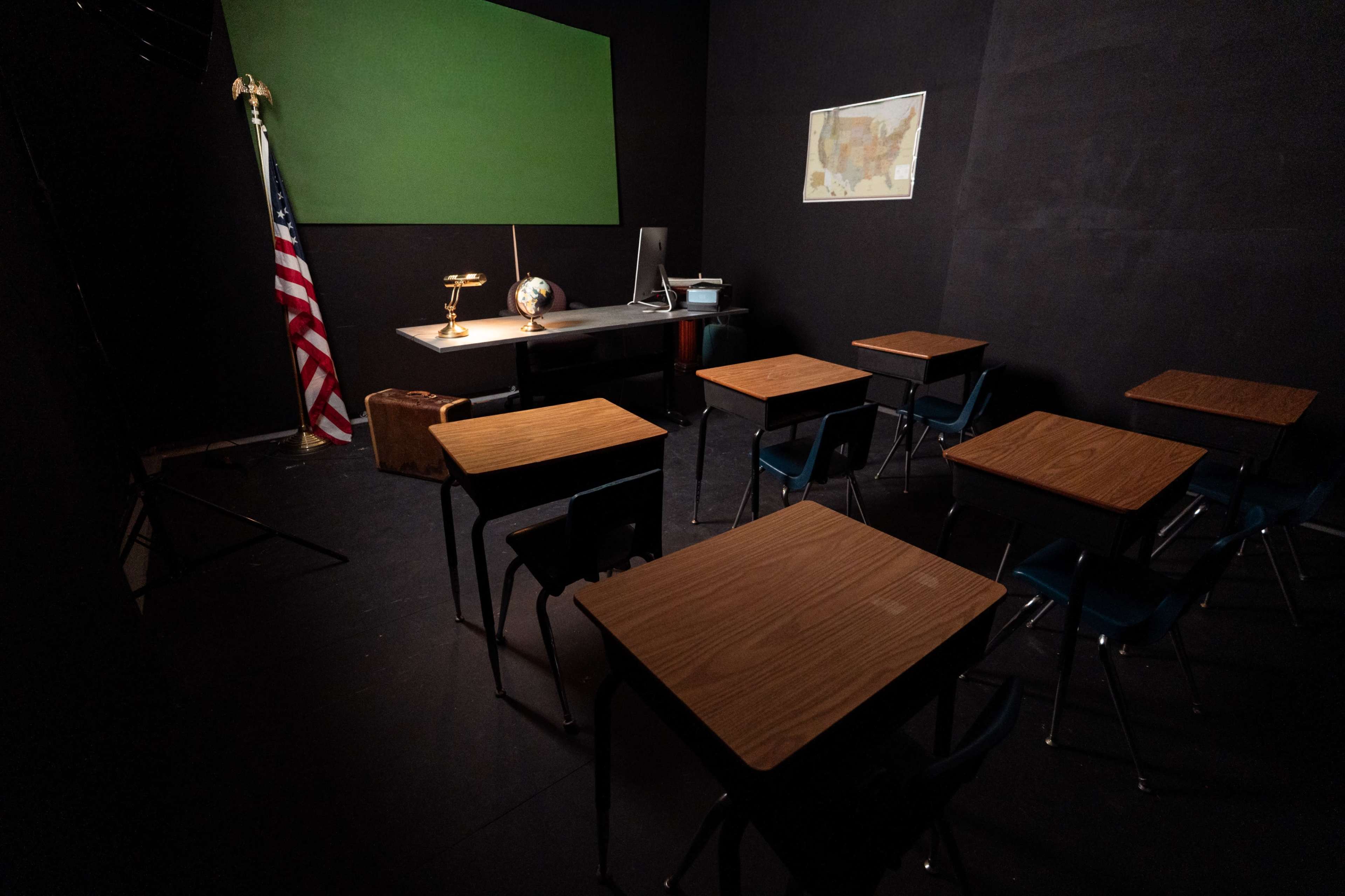 The image shows a classroom setting with empty wooden desks, an American flag, a globe, and a green chalkboard.