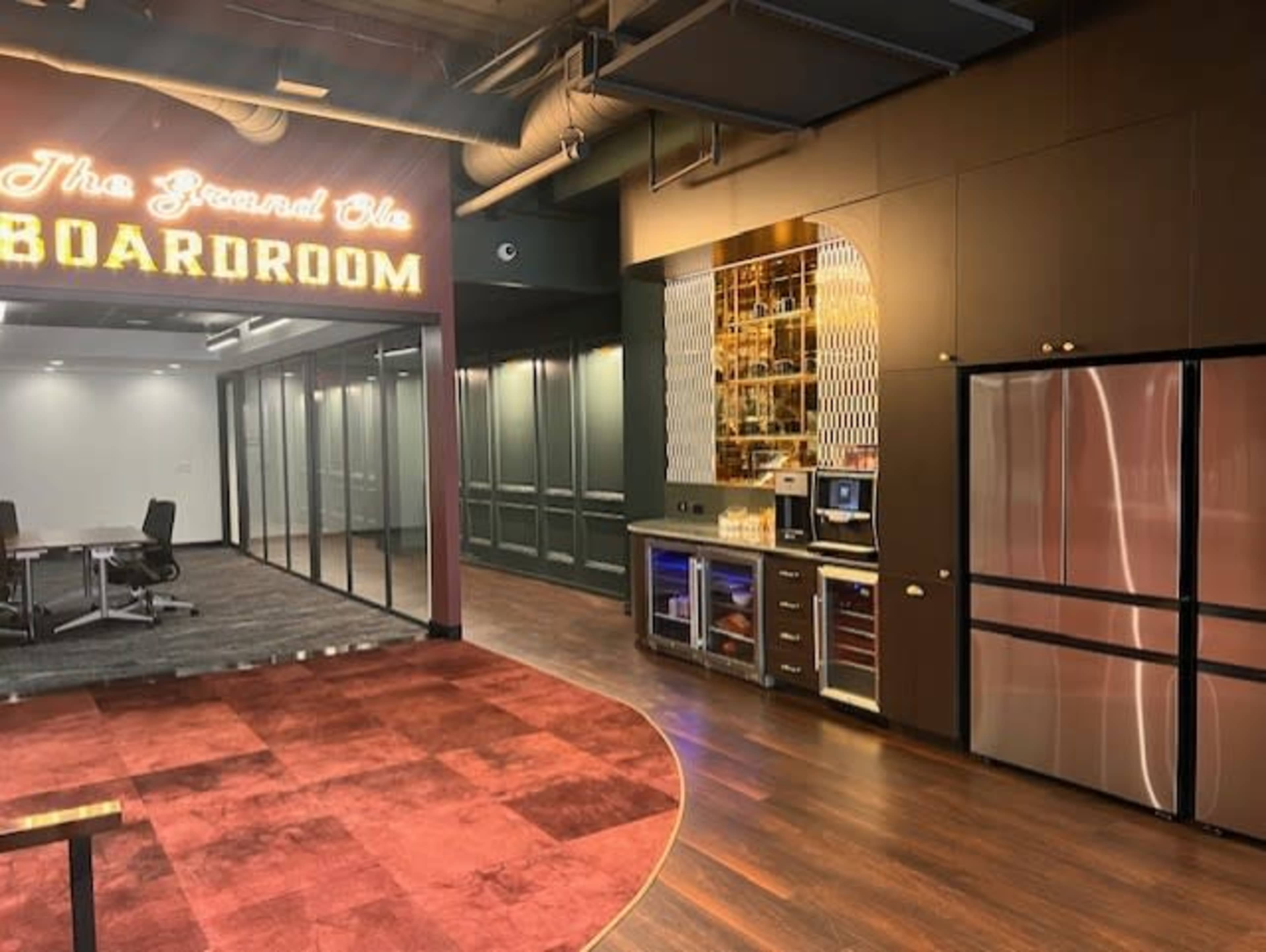 The image shows a modern office space featuring a boardroom entrance with illuminated signage and a kitchenette area that includes a refrigerator, microwave, and bar shelves.