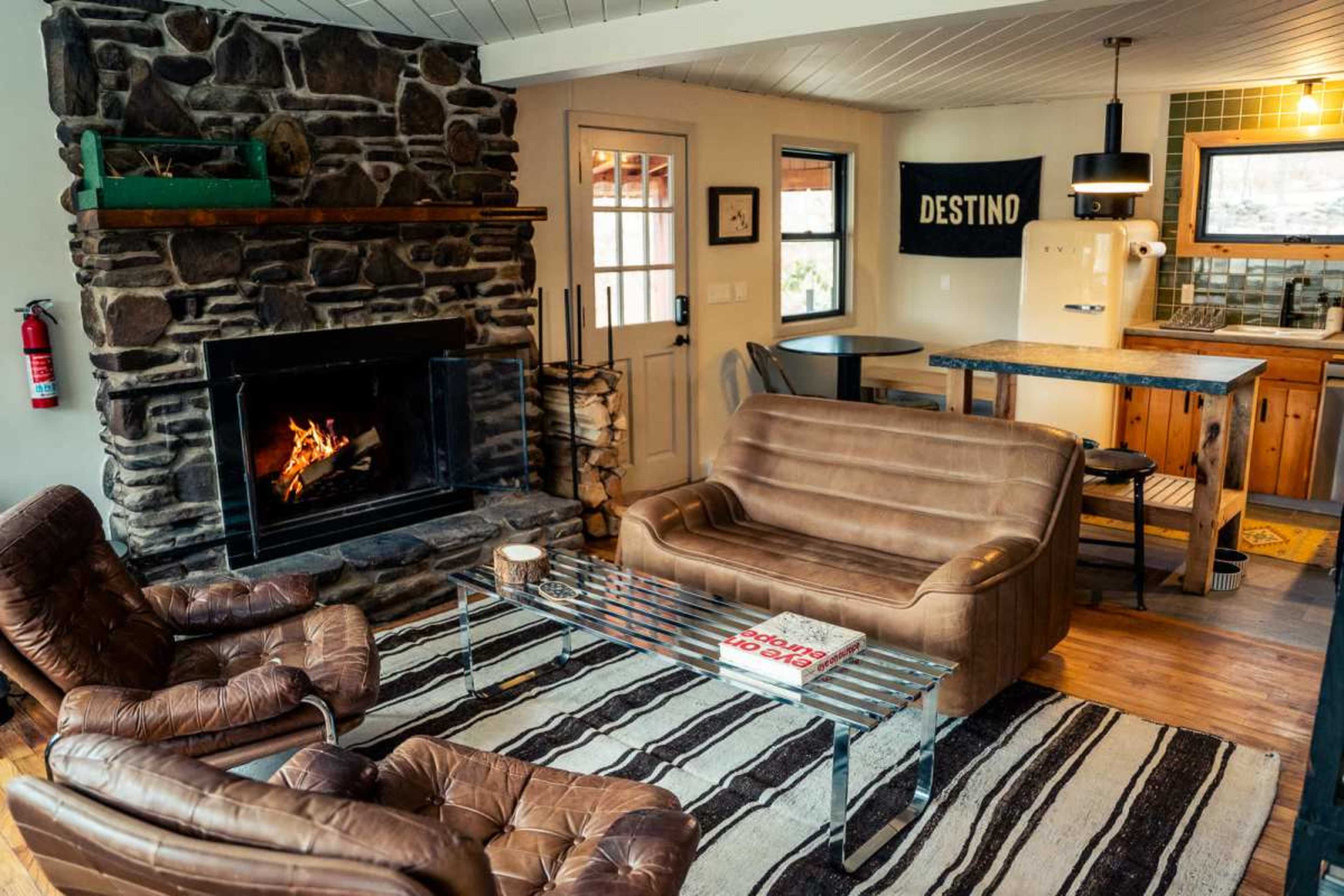 The image shows a cozy living room with a stone fireplace, two brown leather chairs, a glass coffee table, and a view of a kitchen area in the background.