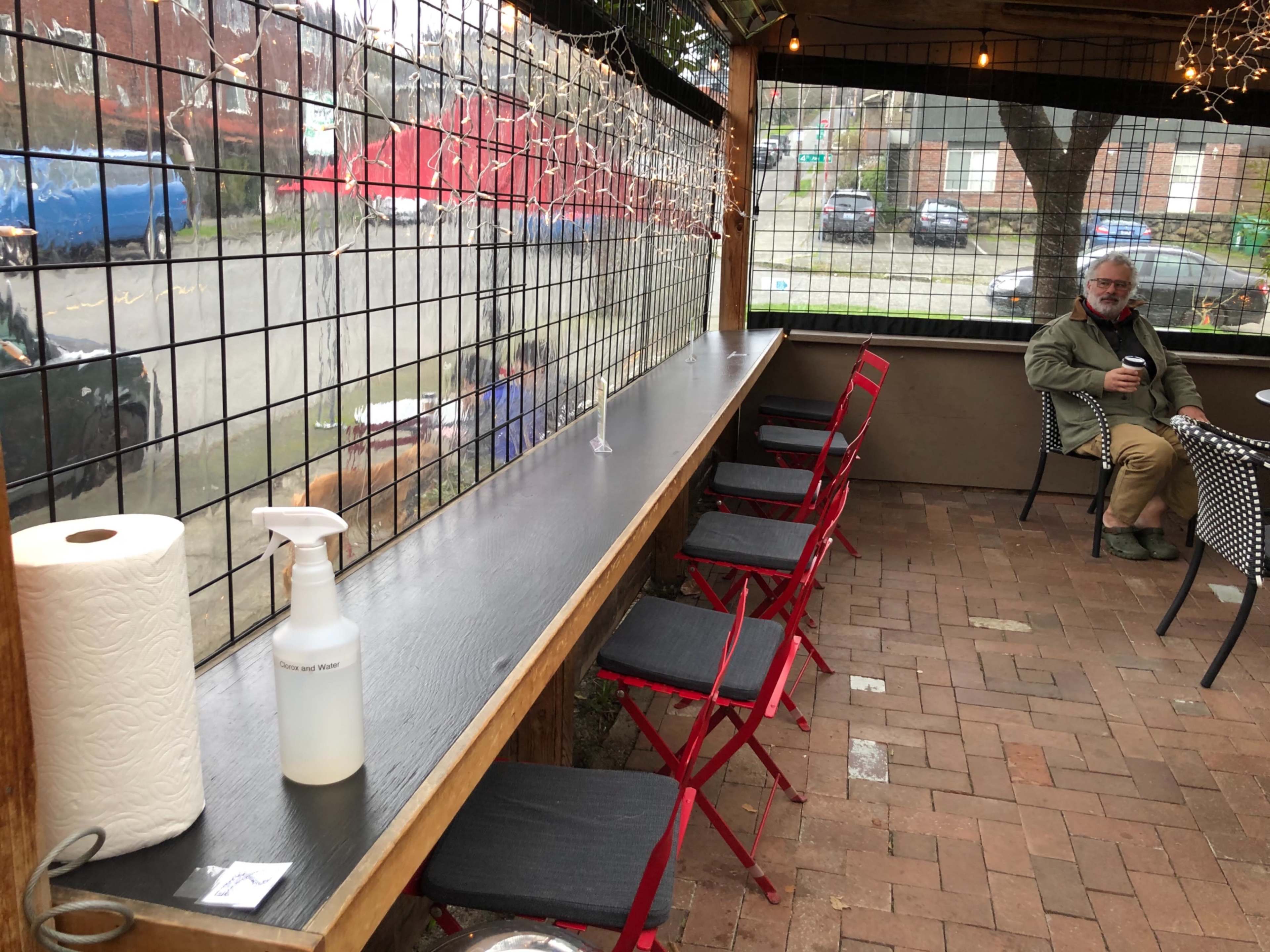 The image shows an outdoor seating area at a café with a long wooden table, red chairs, and a glass barrier, while a man sits nearby holding a cup.