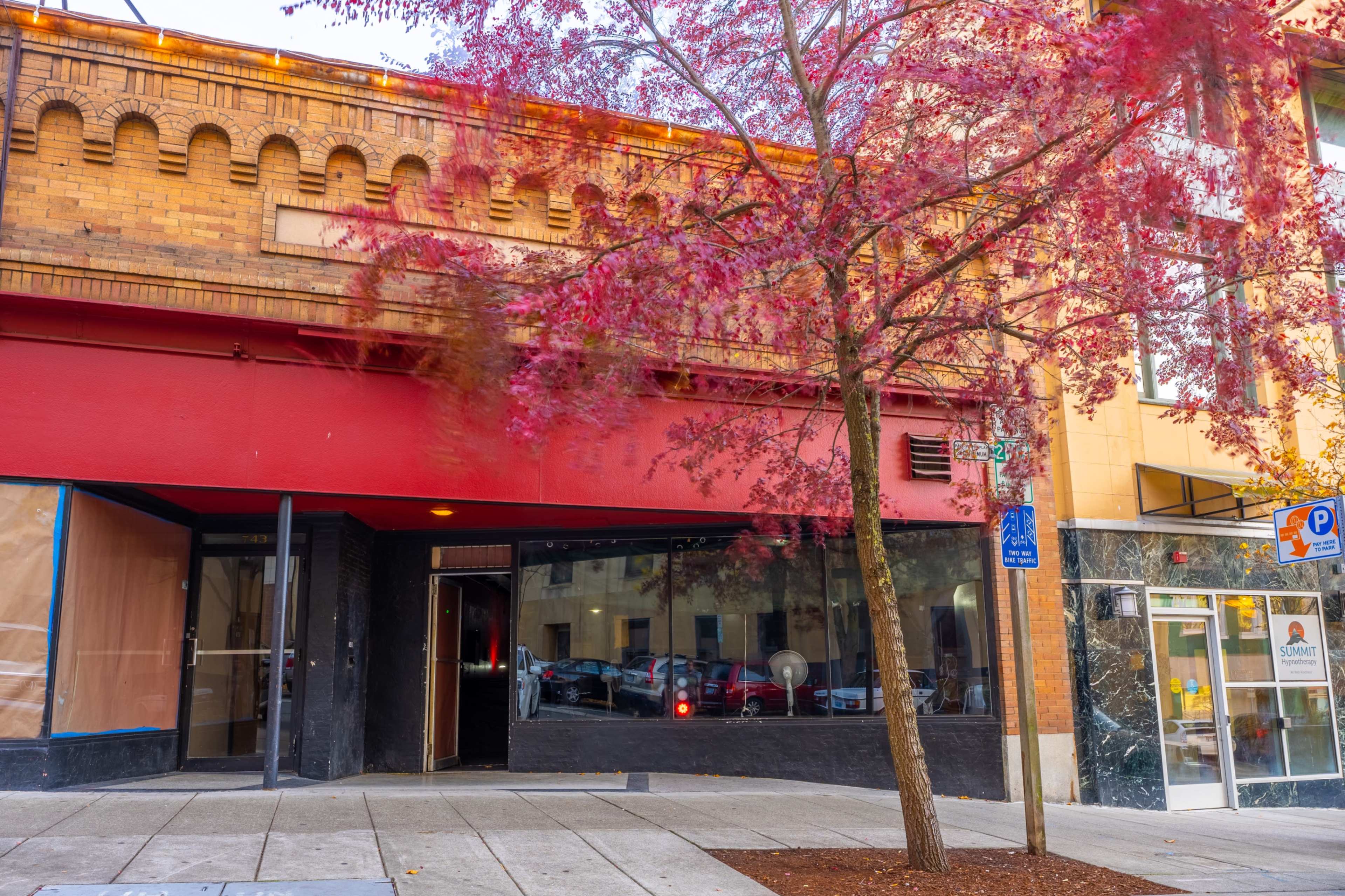 A red-brick building features a partially covered storefront and a tree with red leaves in front, alongside a contrasting glass structure.