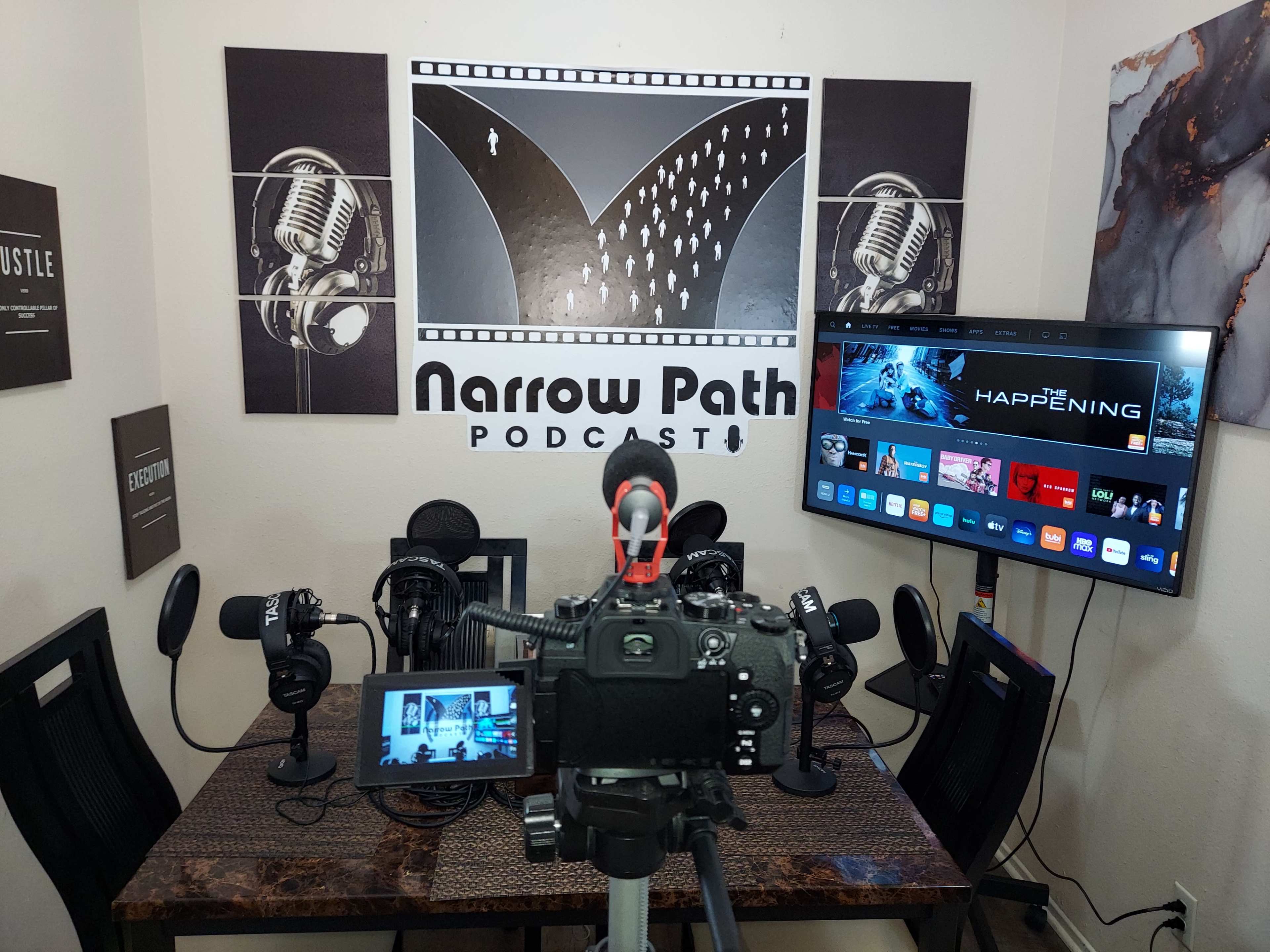 A camera is set up on a table facing a podcast studio with microphones and a large screen displaying content related to the "Narrow Path Podcast."