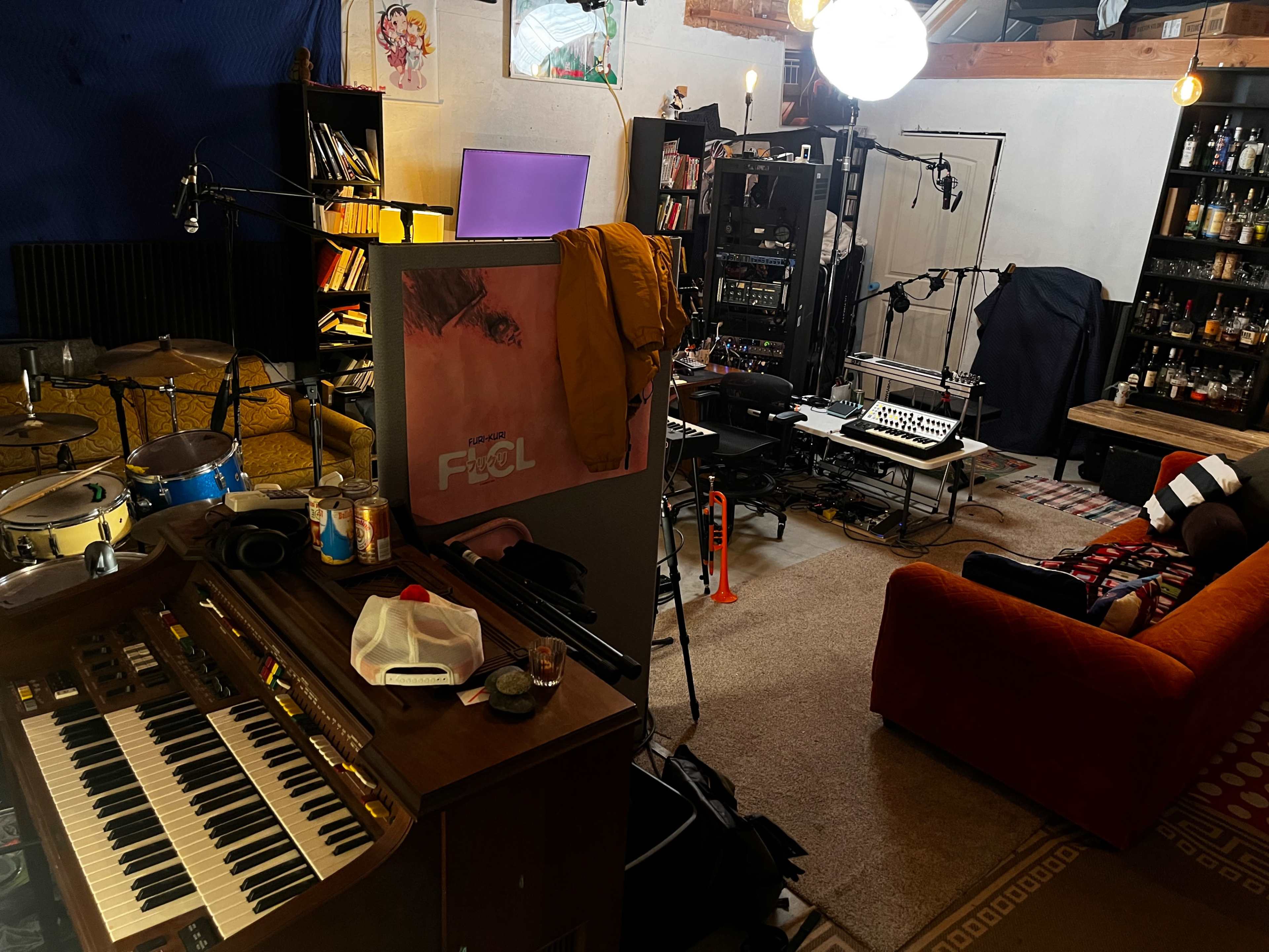 A cluttered music studio with a keyboard, drum set, and various recording equipment surrounded by a couch and shelves filled with bottles.