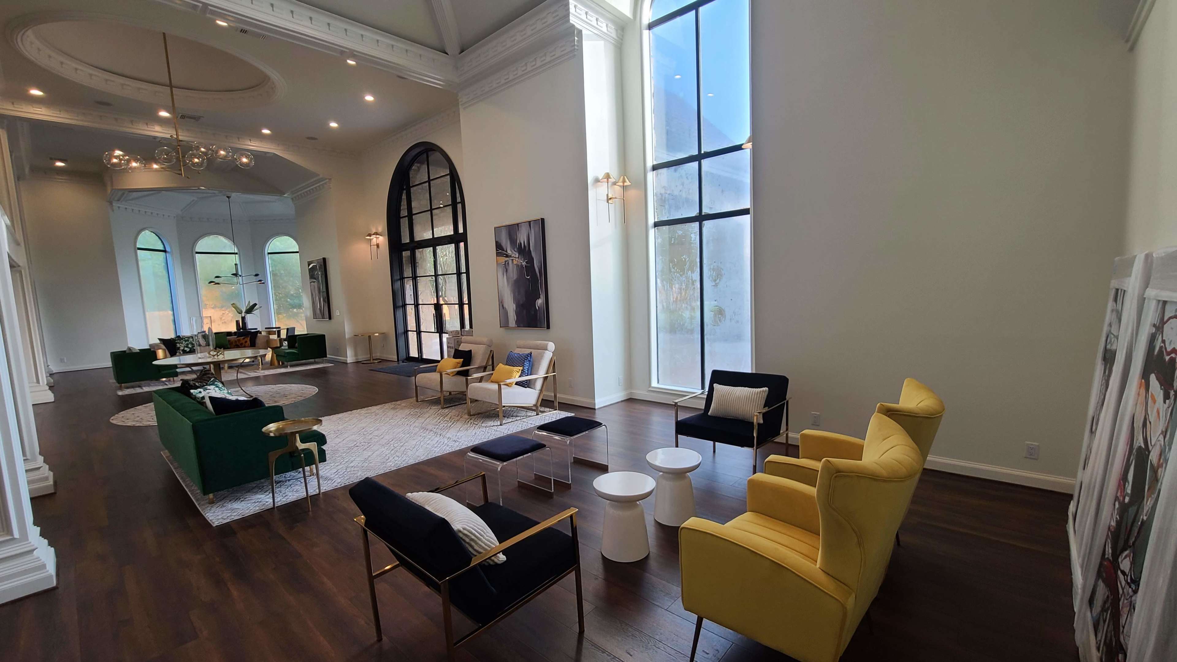 The image shows a spacious, modern living area with large windows, featuring a mix of seating options in different colors and styles.