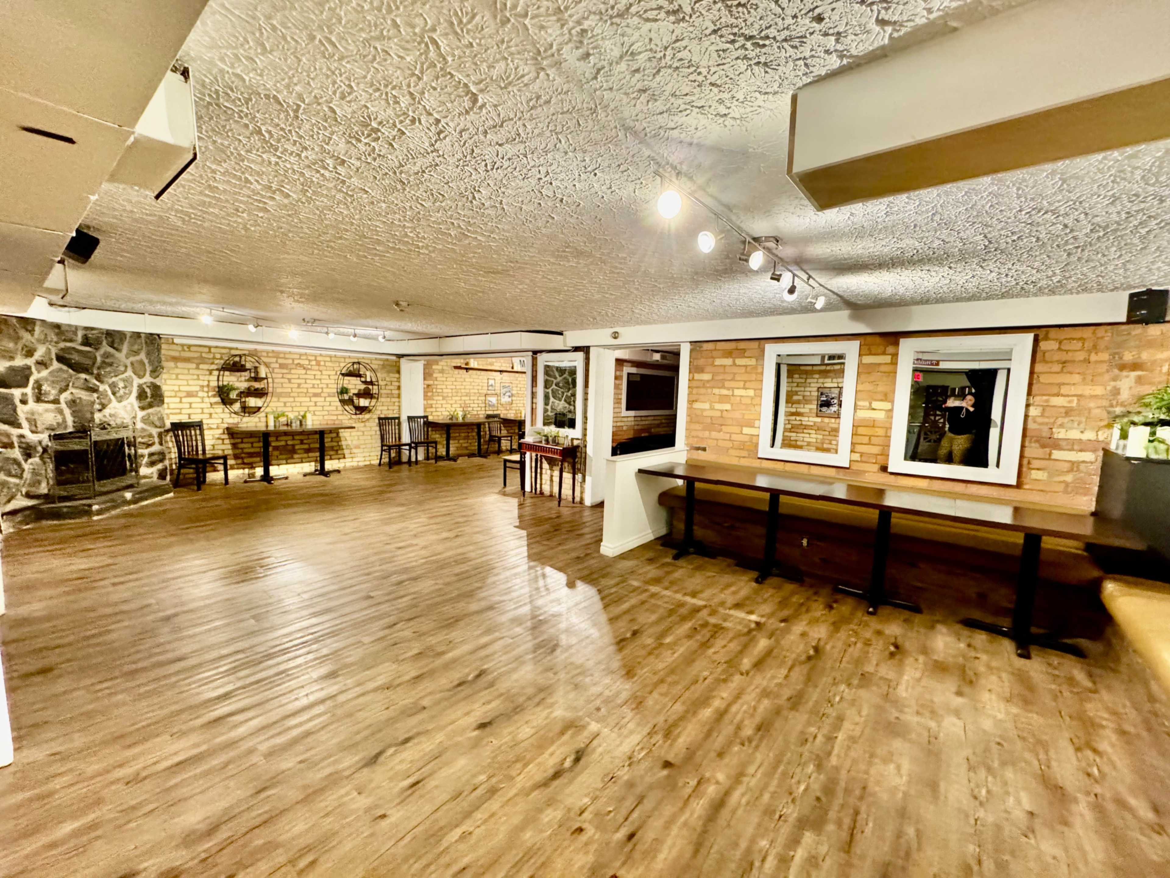 A spacious, well-lit room with wooden floors, brick walls, and several tables and chairs arranged around the area.