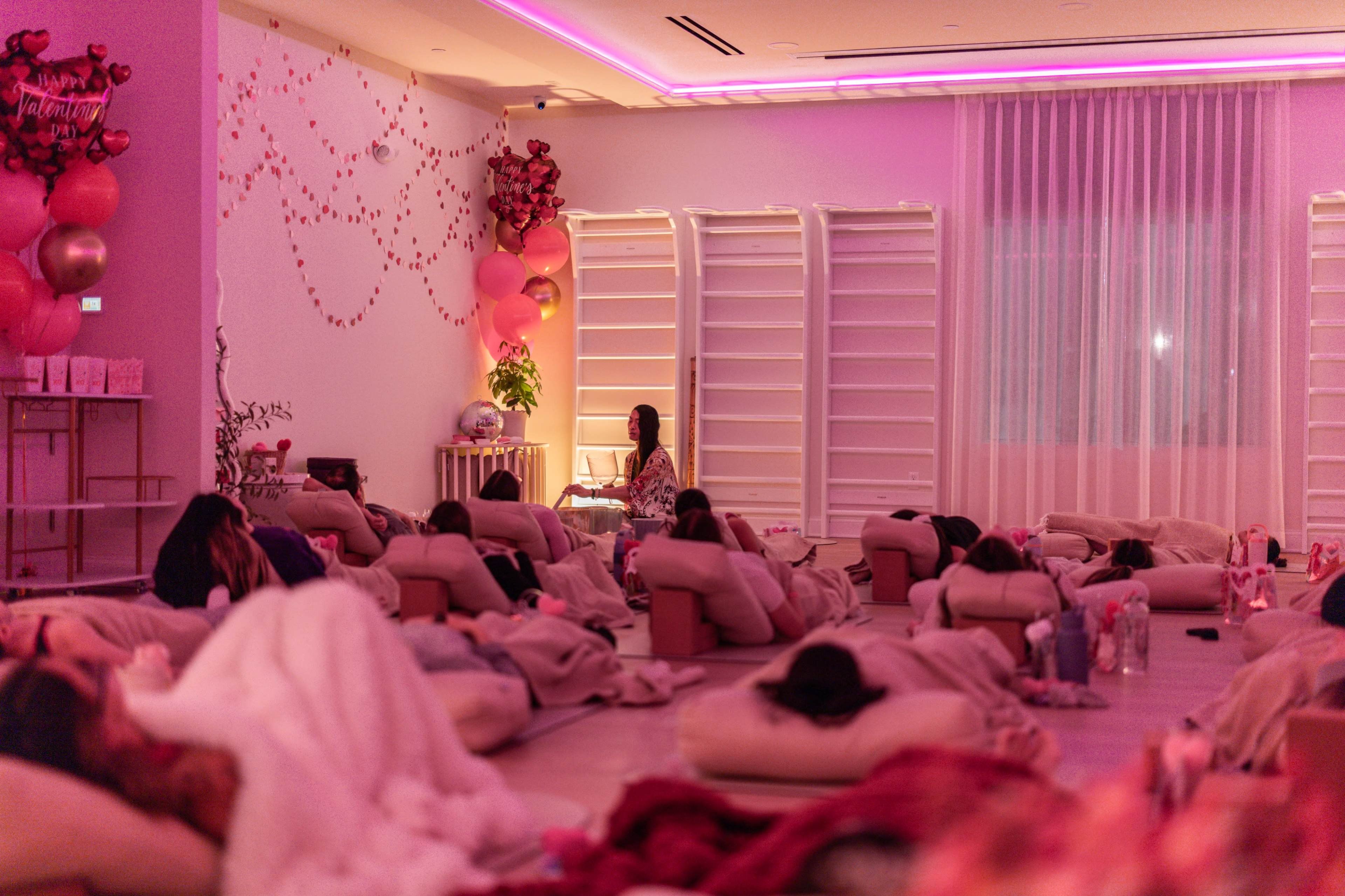The image shows a dimly lit room with people lounging on cushioned mats, surrounded by pink decorations and a presentation setup at the front.