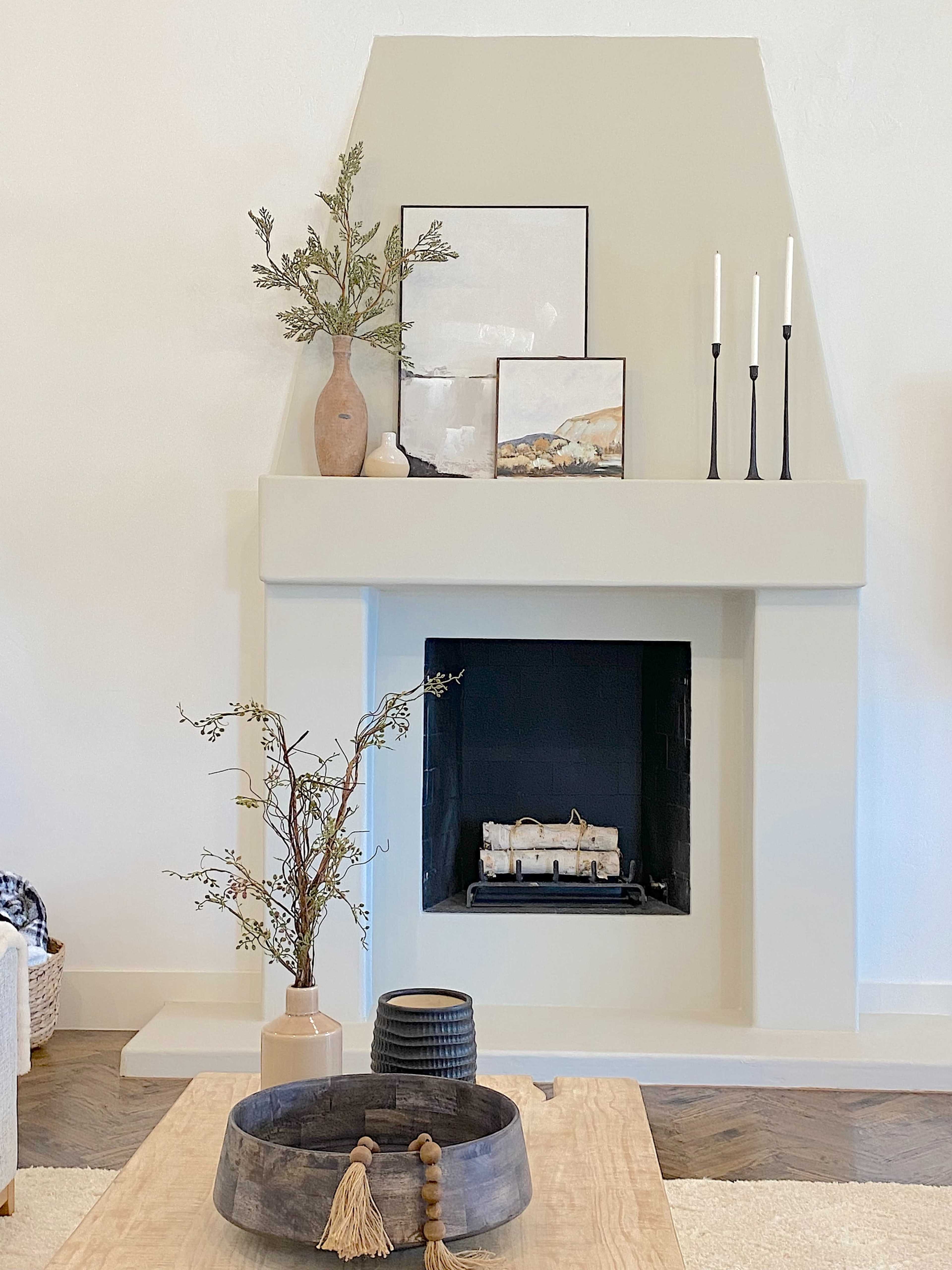 A minimalist living room features a light-colored fireplace with a decorative vase, artwork, and candles arranged on the mantel, alongside a wooden coffee table with a circular tray.