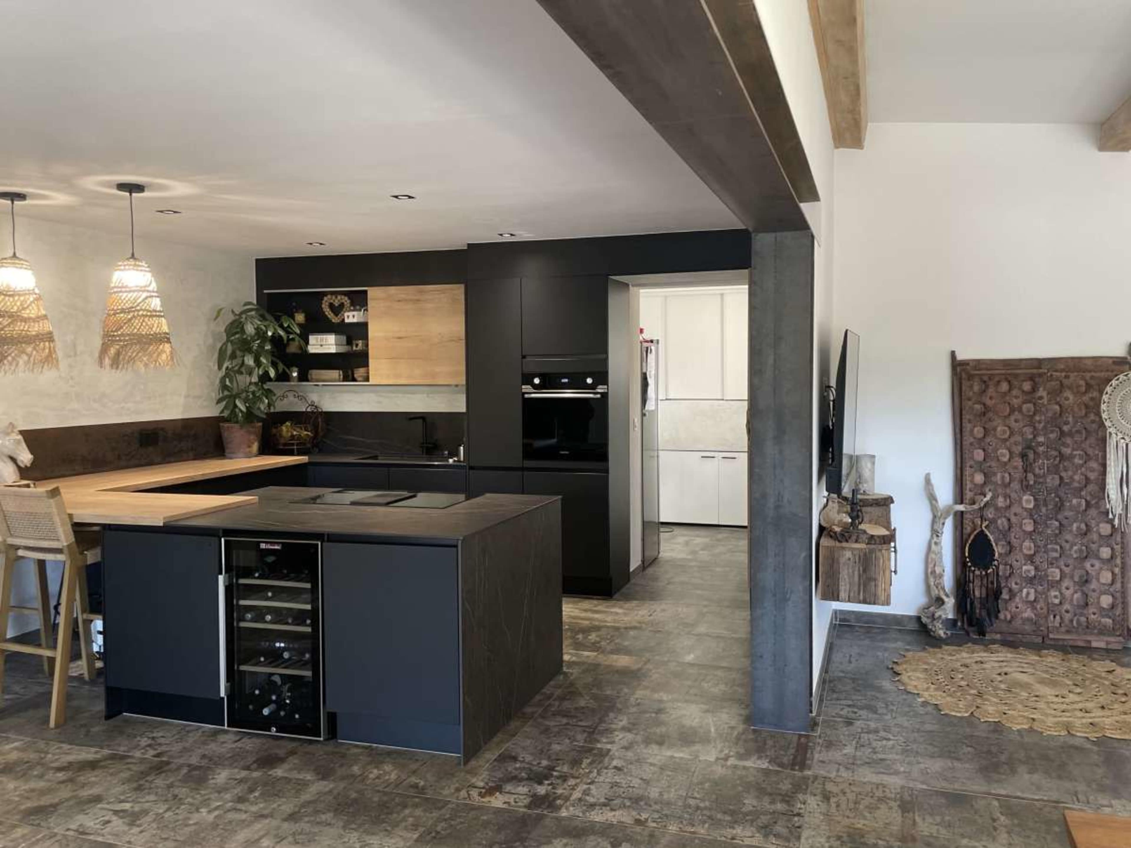 The image shows a modern kitchen featuring a central island with a cooktop, black cabinetry, and wooden accents, alongside a dining area with wicker chairs.