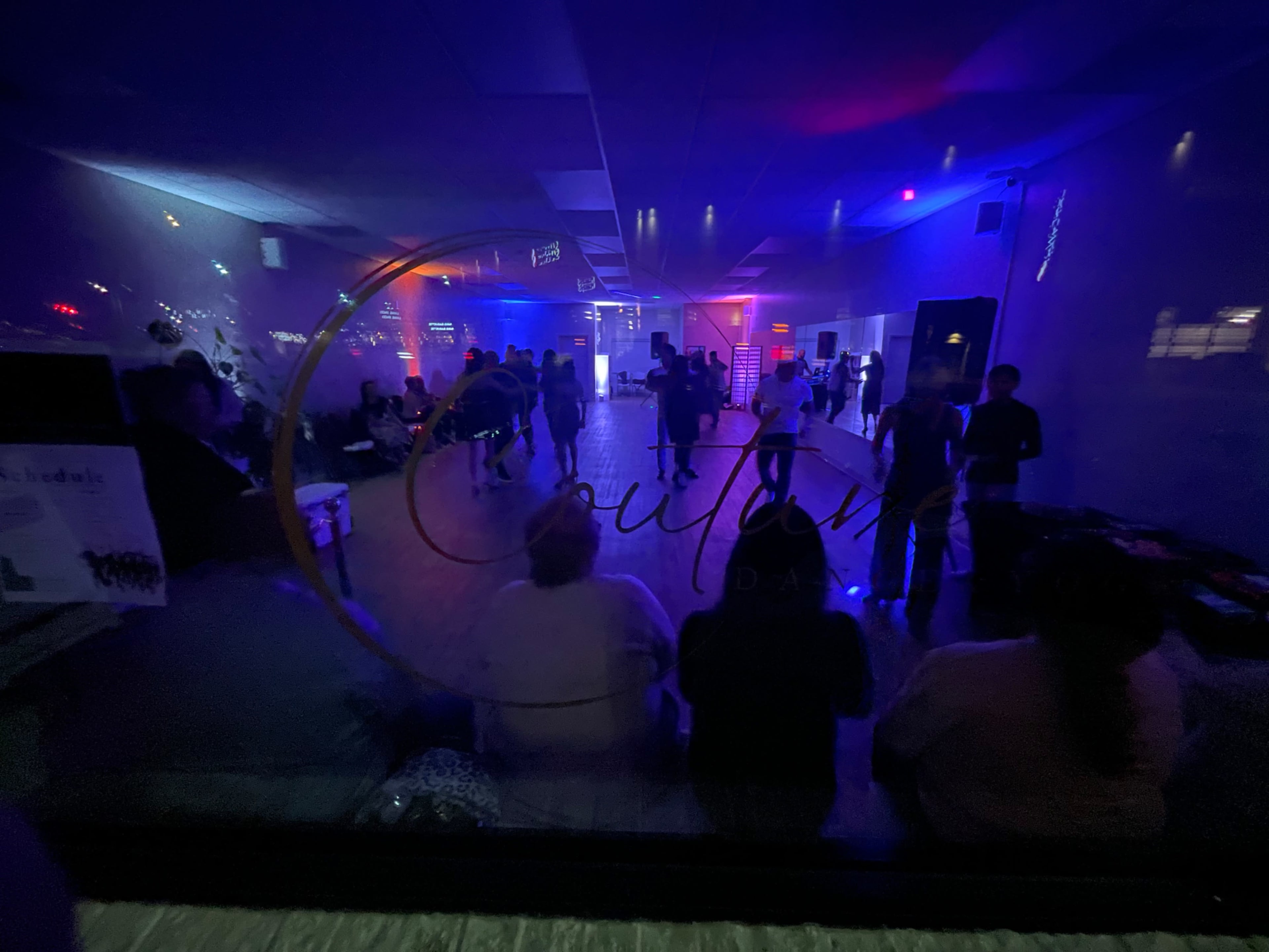 A dimly lit dance hall with people interacting and colorful lights illuminating the space.