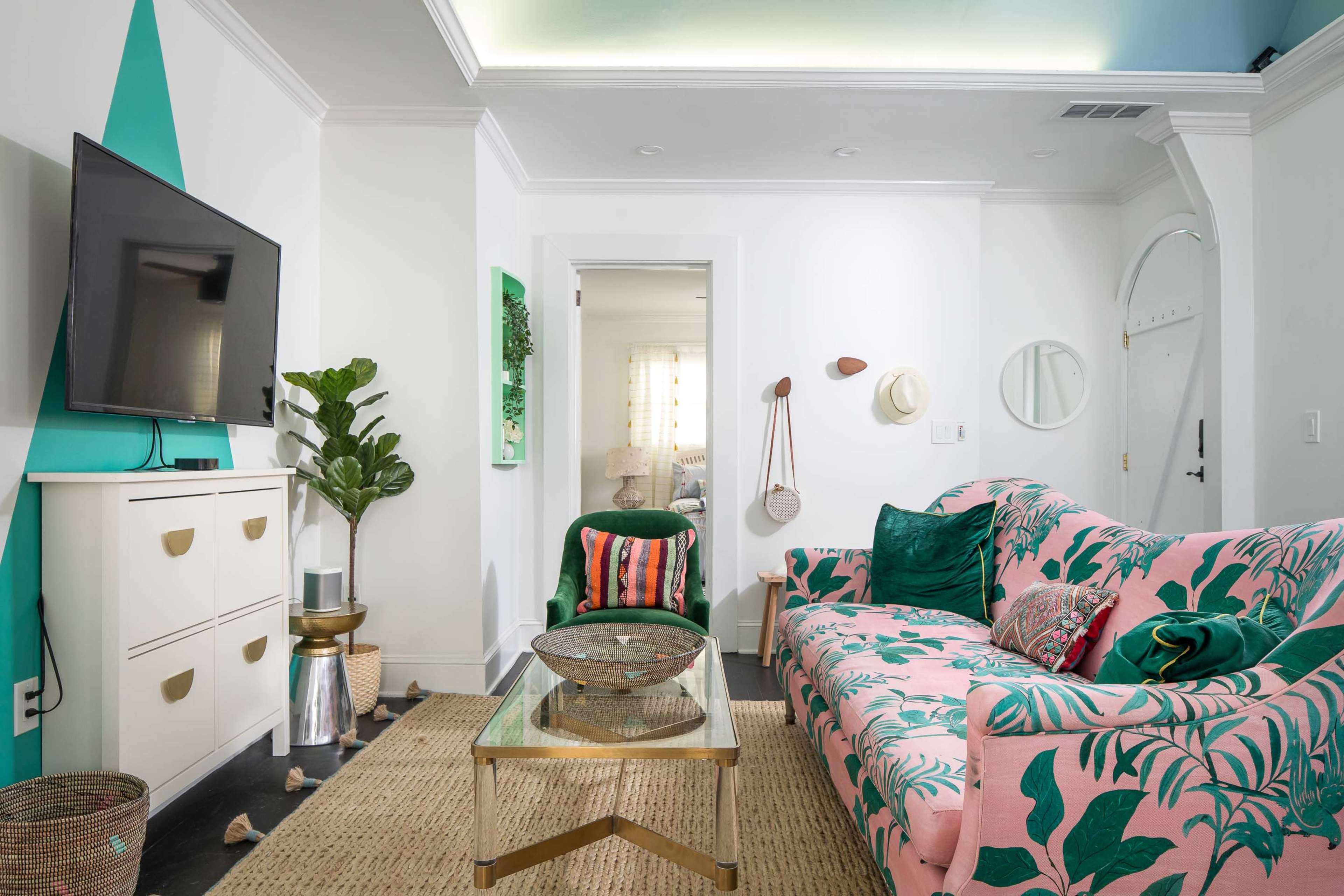 The room features a pink, leafy-patterned sofa, a green armchair, a glass coffee table, and a modern television against a white wall.