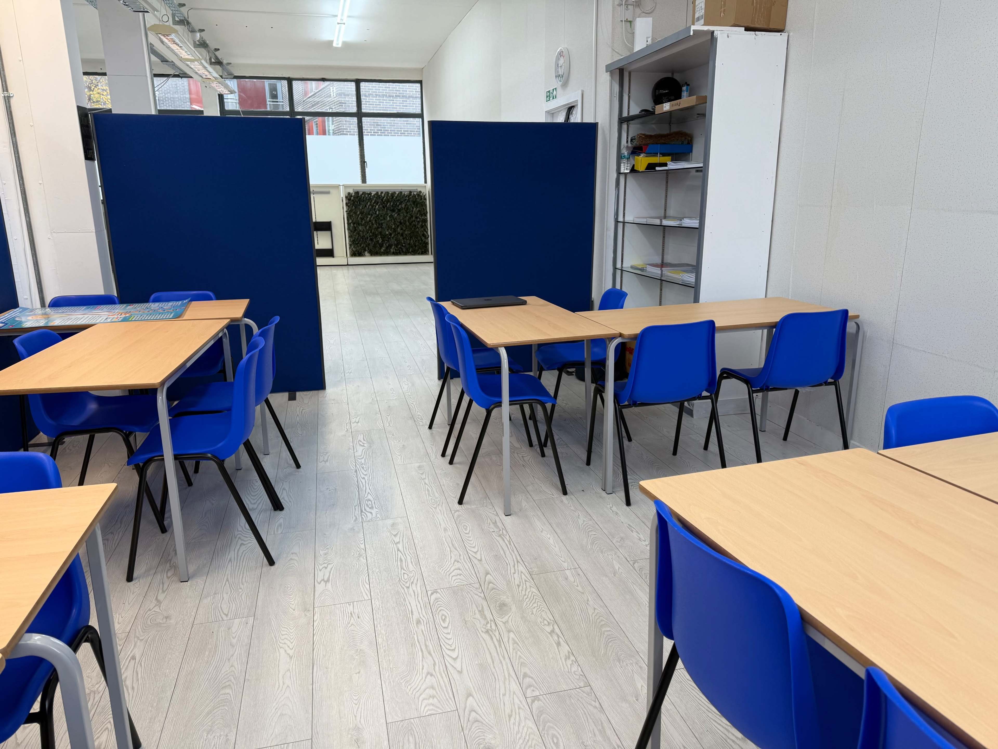 The image shows a classroom with several blue tables and chairs arranged around a mostly empty space, featuring blue panels along one side.