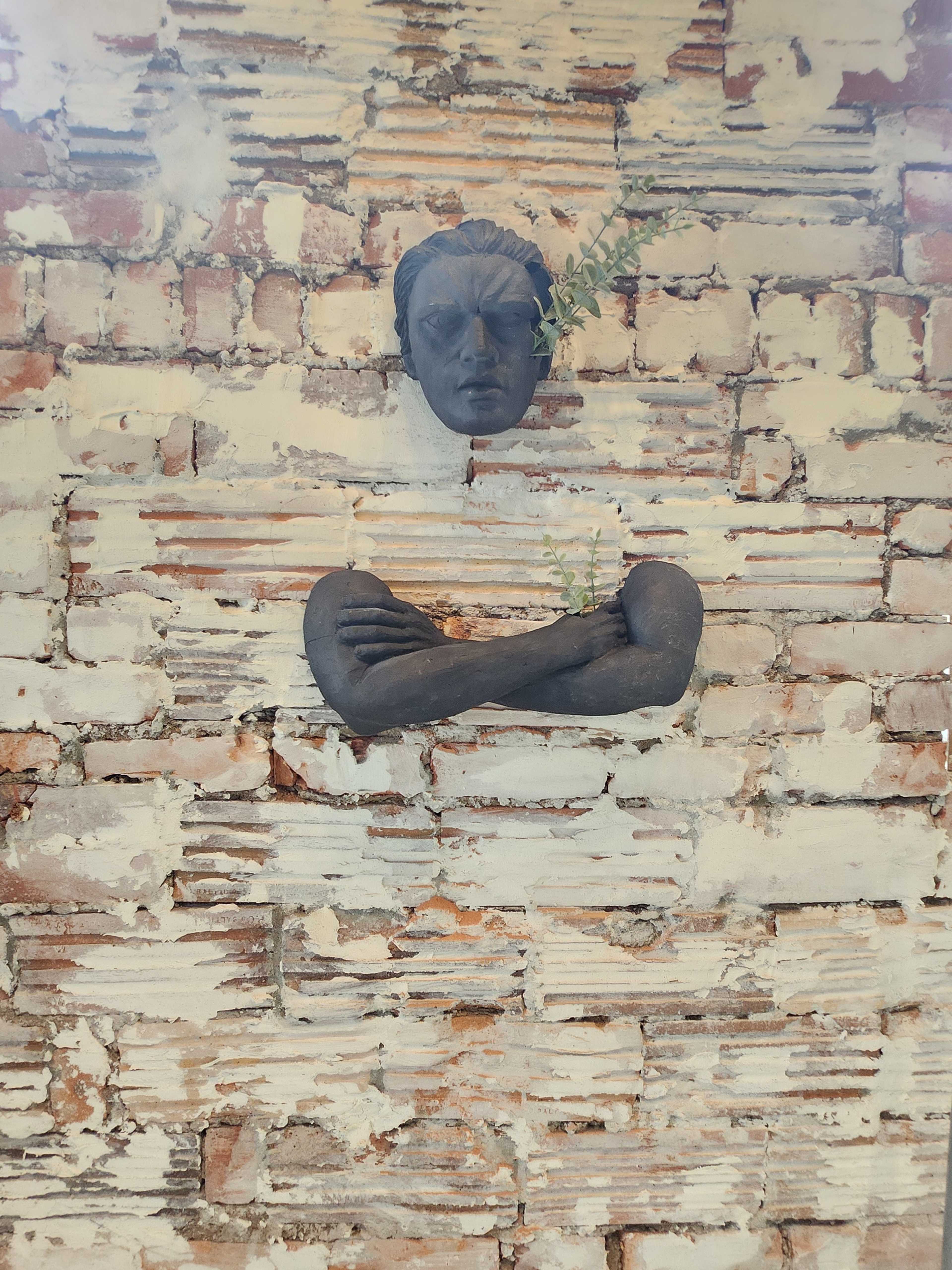 A sculpted head and arms are mounted against a weathered brick wall, with a small plant growing between the figure's arms.