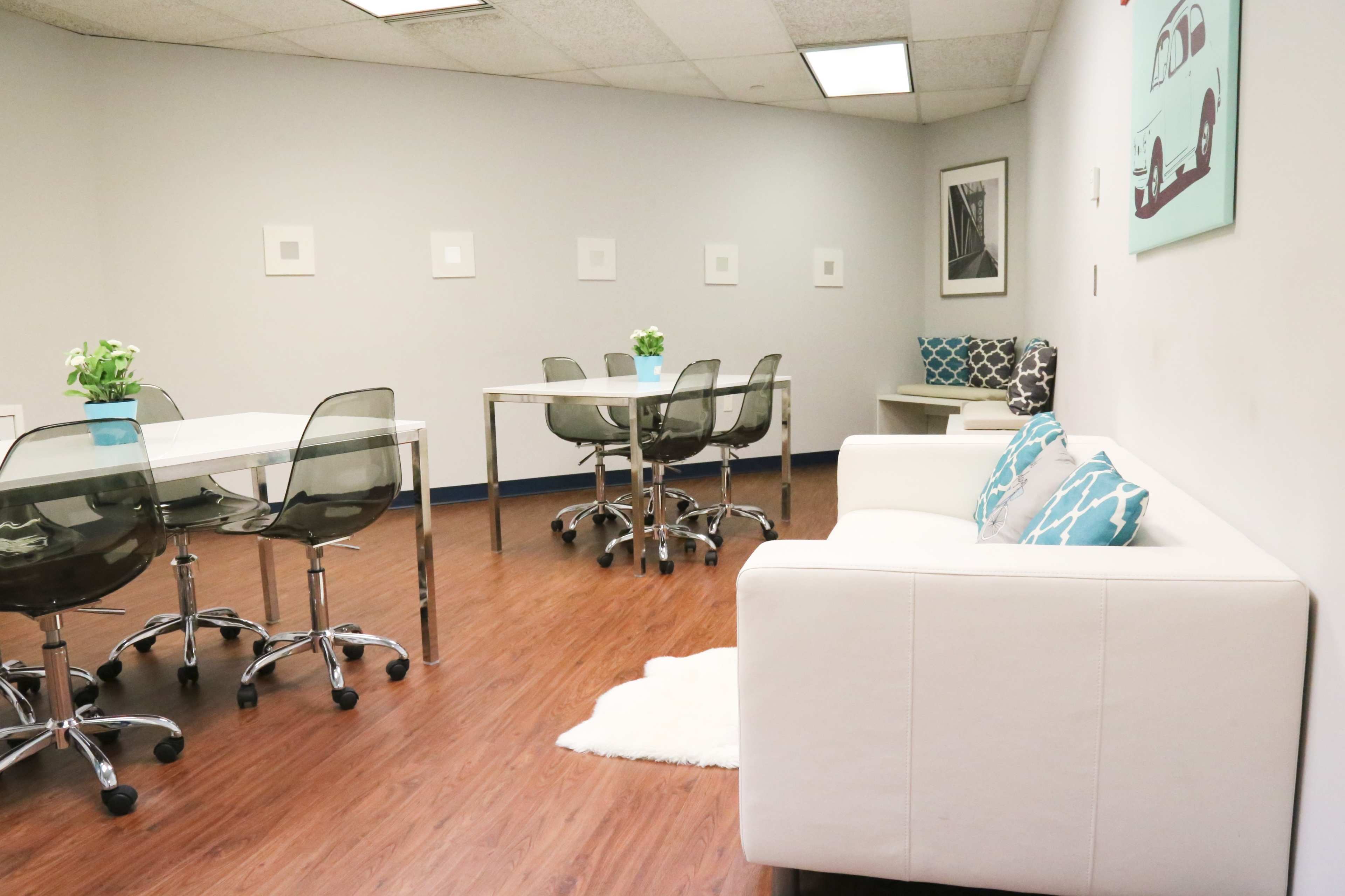 A modern office space with white walls, a light wooden floor, a white sofa, and several glass tables surrounded by transparent chairs.