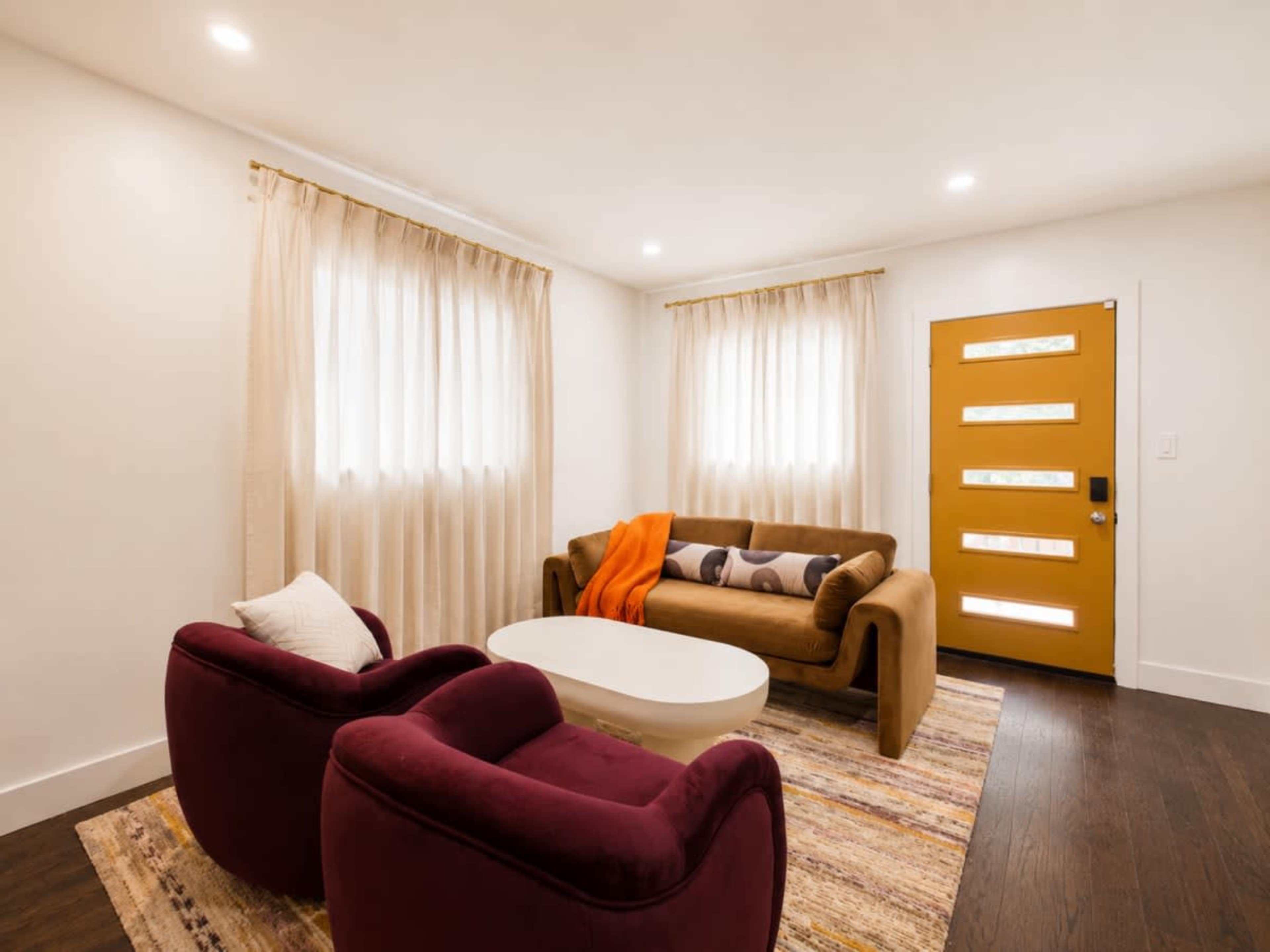 The image shows a modern living room featuring a brown sofa, two burgundy chairs, a white coffee table, and natural light streaming through sheer curtains.