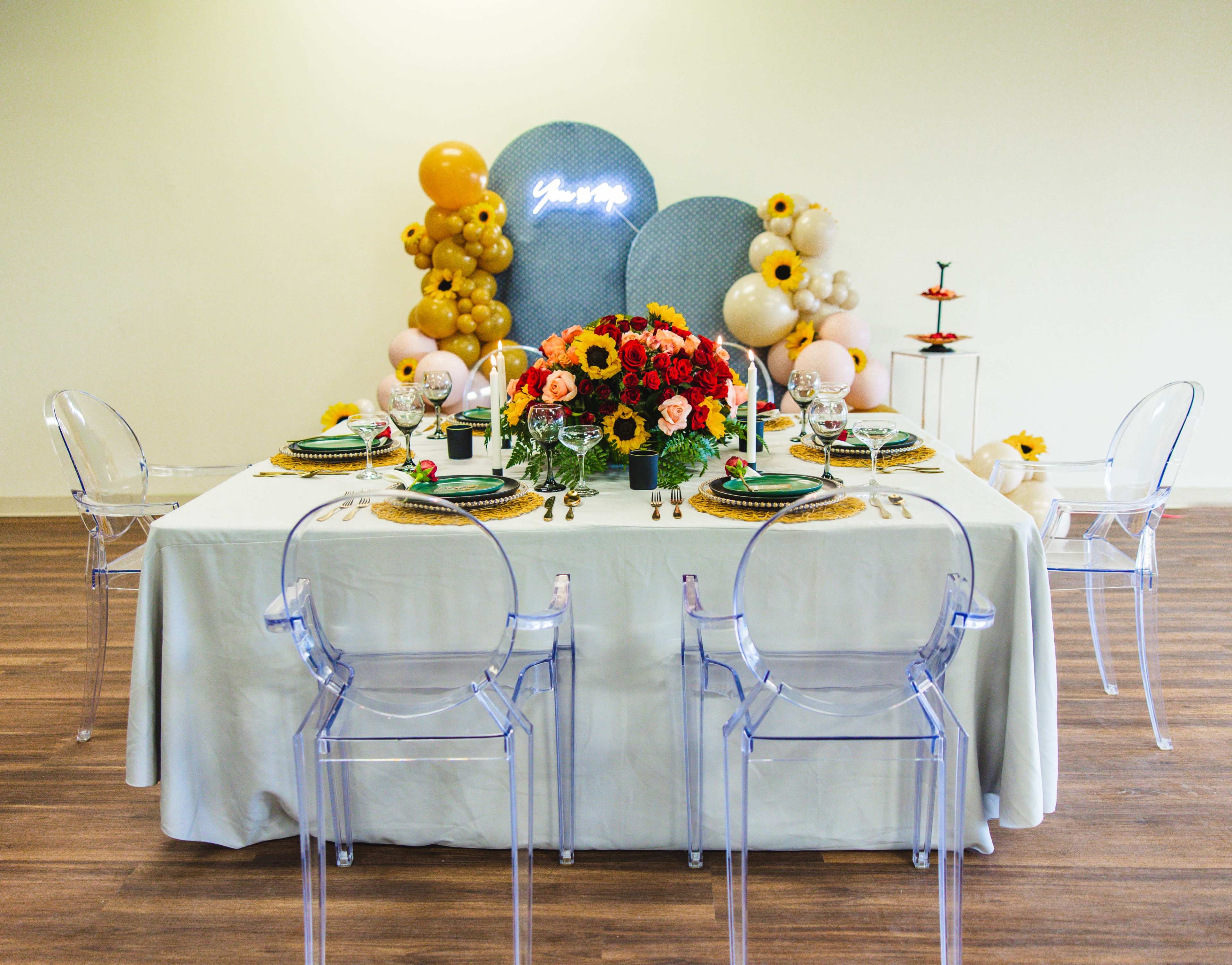 A decorated banquet table with floral arrangements, elegant tableware, and transparent chairs is set up in a bright room.
