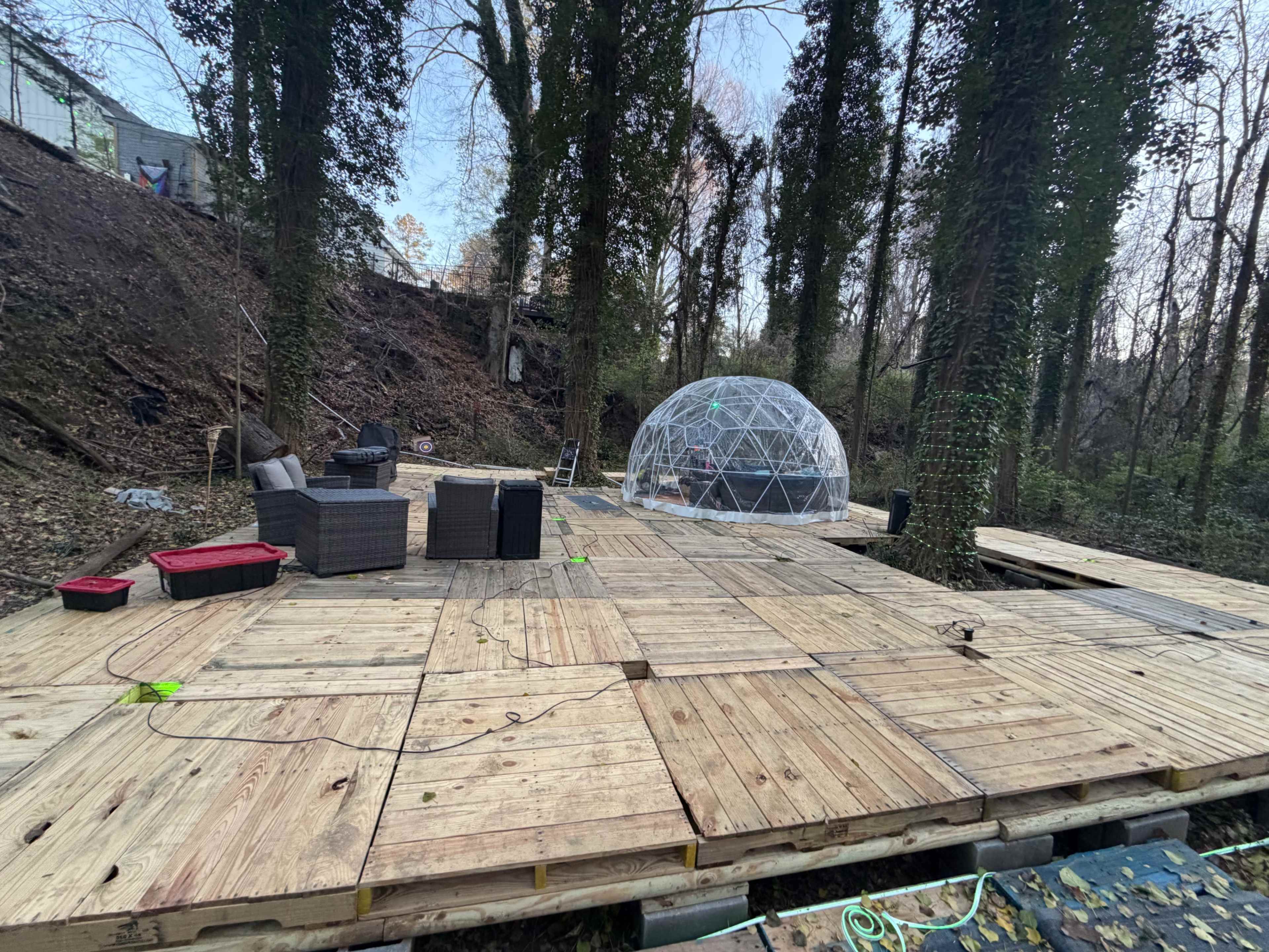 A wooden deck with furniture is set up in a wooded area, featuring a geodesic dome in the background.