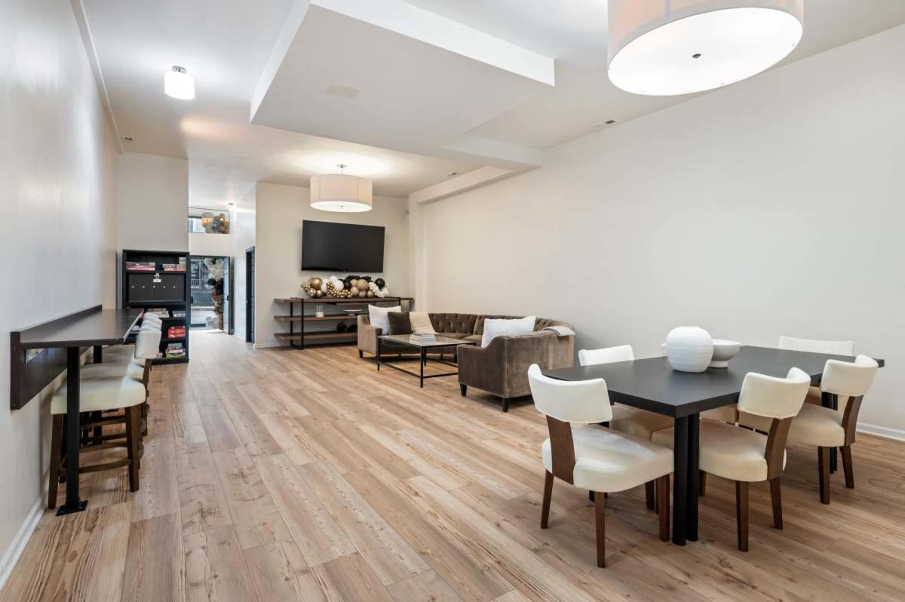 The image shows a light-filled modern interior featuring a dining area with a black table and white chairs, a brown sofa, and a television mounted on the wall.