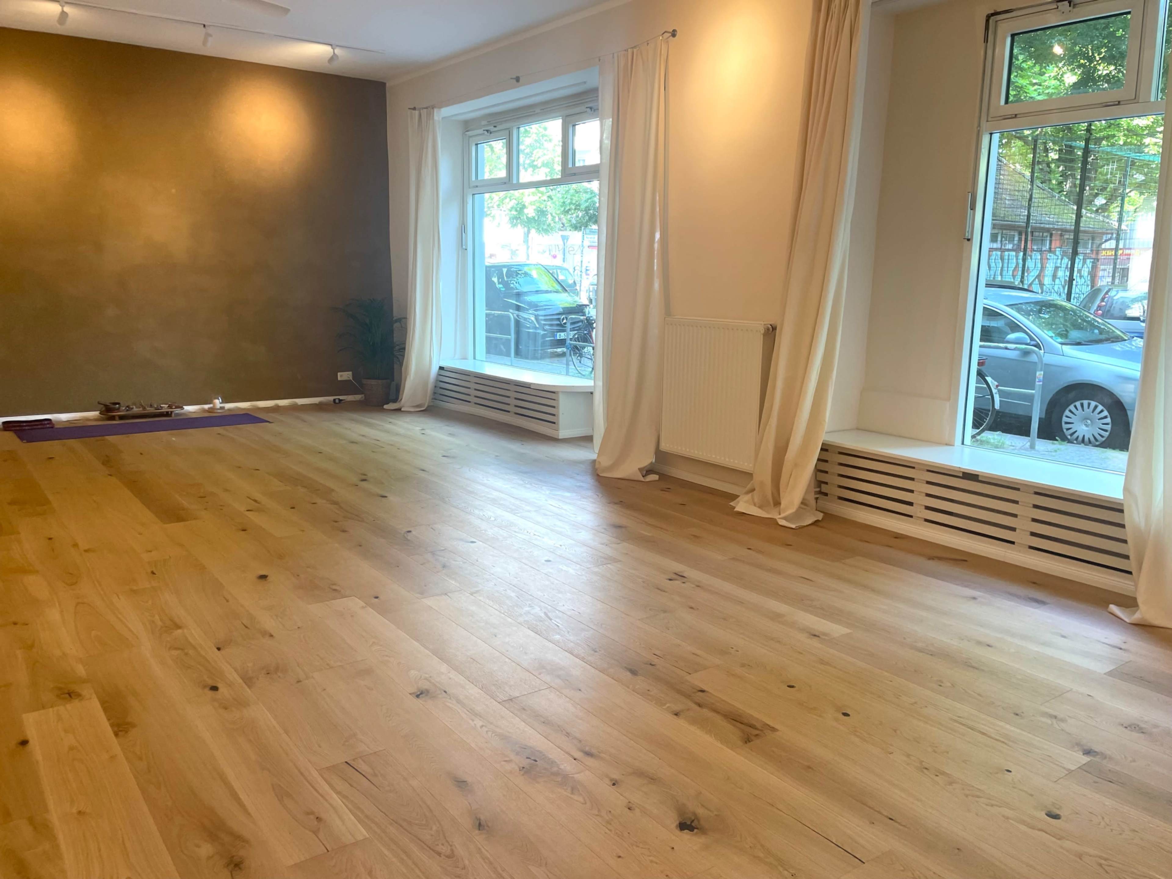 The image shows a spacious, well-lit room with wooden flooring, large windows, and minimal furnishings.