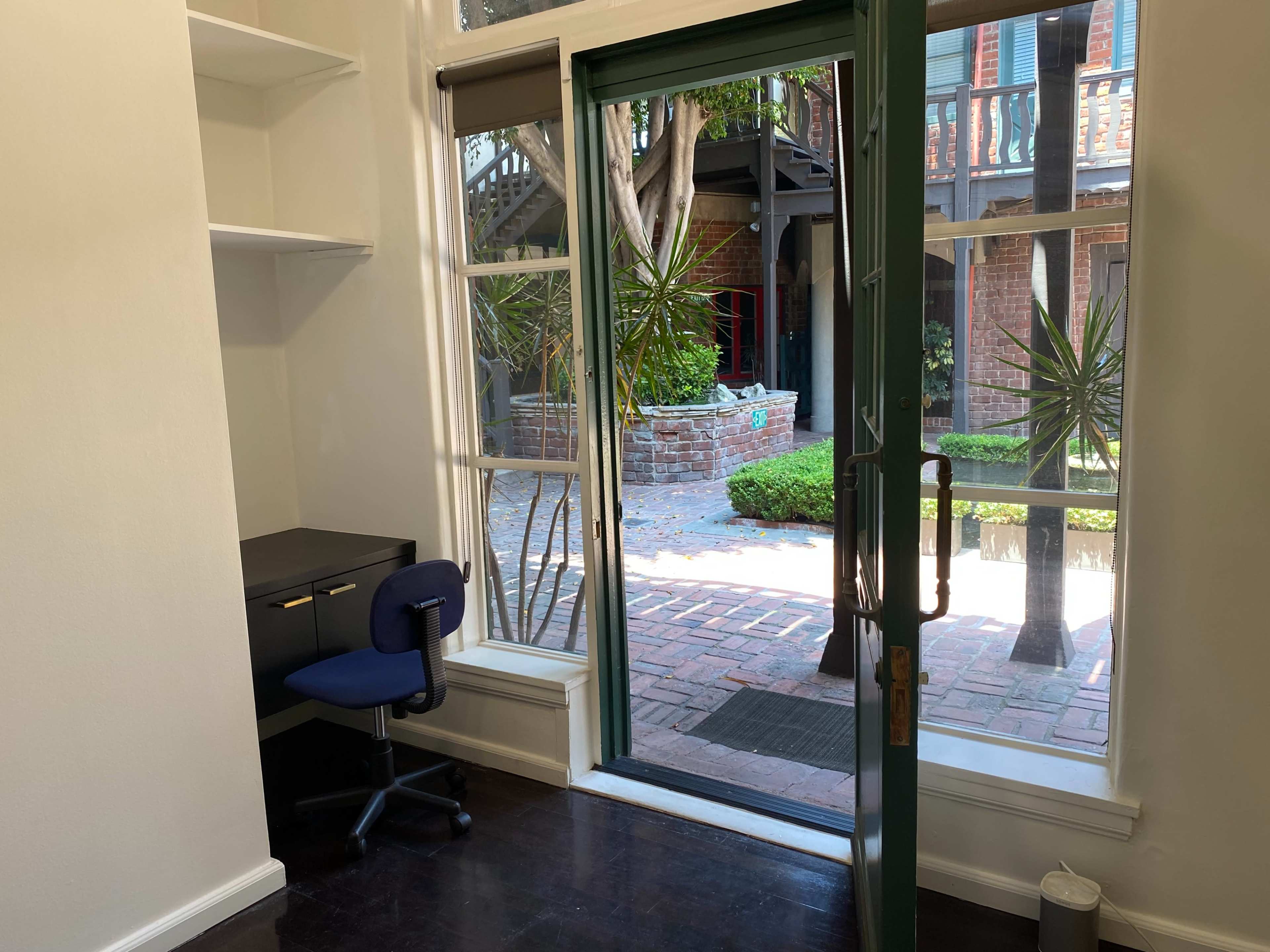 The image shows a small, tidy room with a dark chair and desk positioned near a glass door leading to a courtyard with greenery and brick pathways.