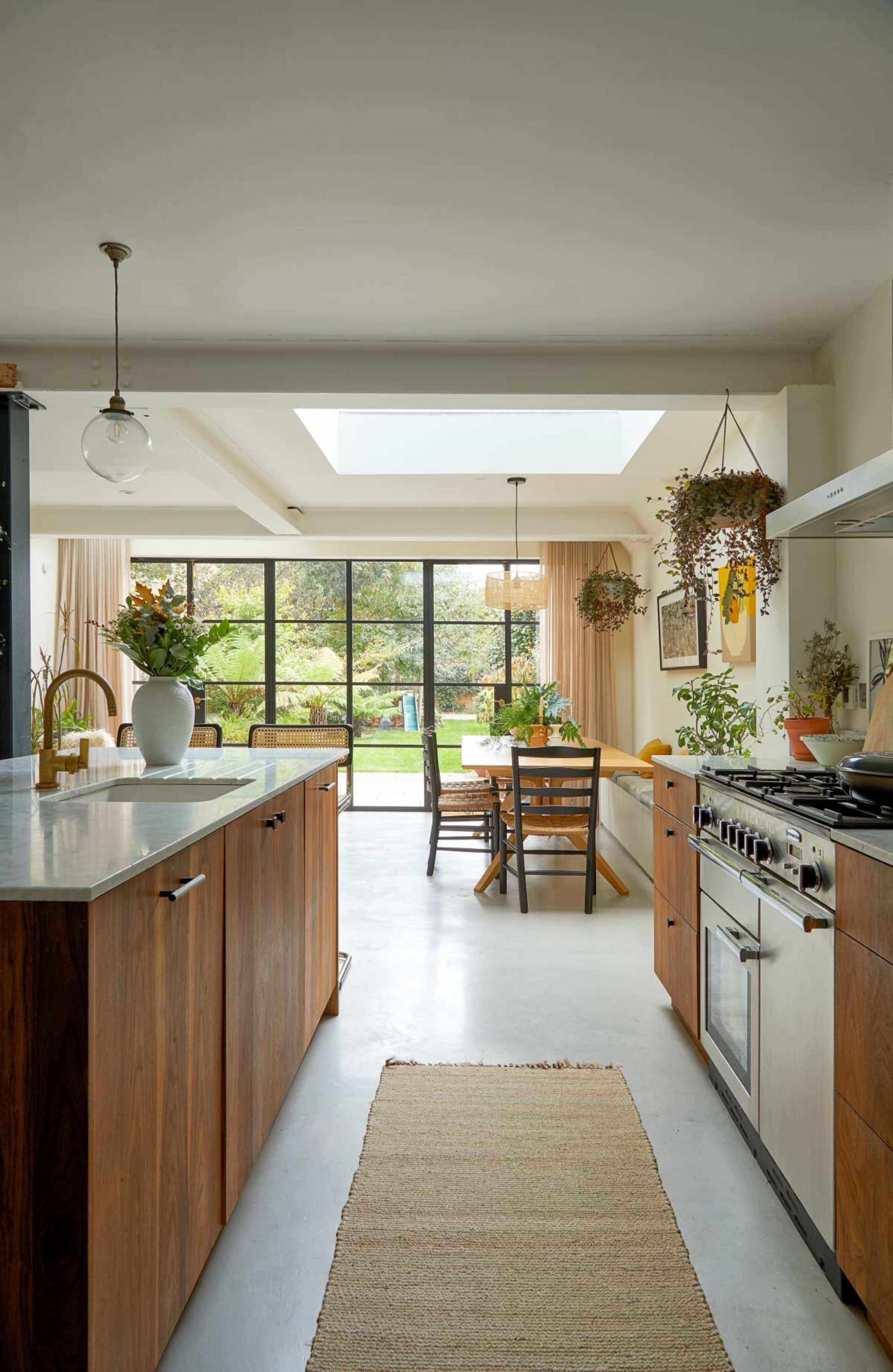 The image shows a modern kitchen featuring wooden cabinetry, stainless steel appliances, and a dining area with a table and chairs, illuminated by natural light from a skylight.