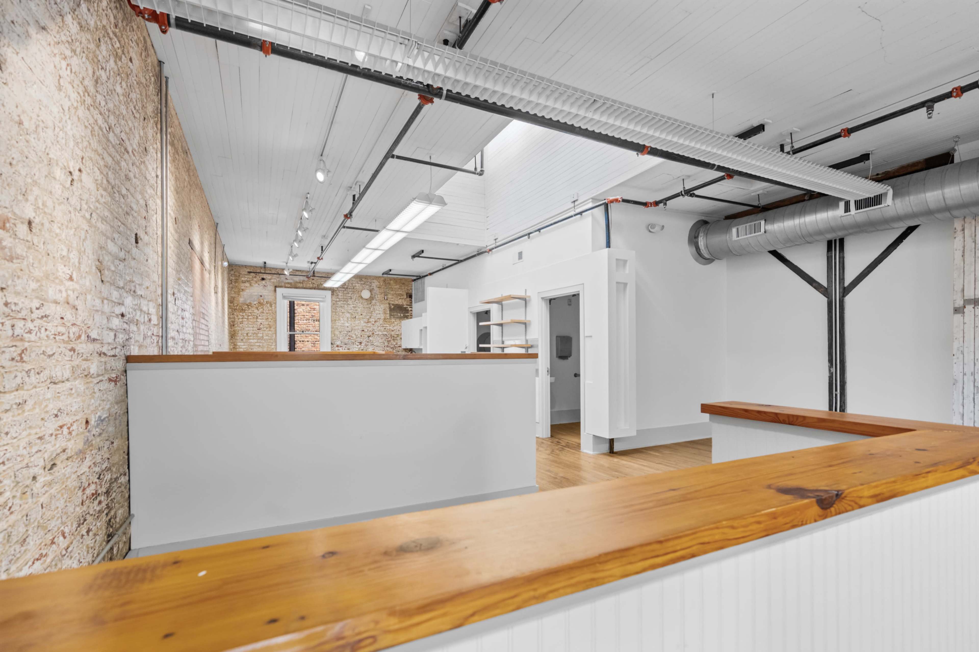 The image shows a spacious, modern interior with exposed brick walls, wooden countertops, and open areas, featuring white walls and sleek lighting.