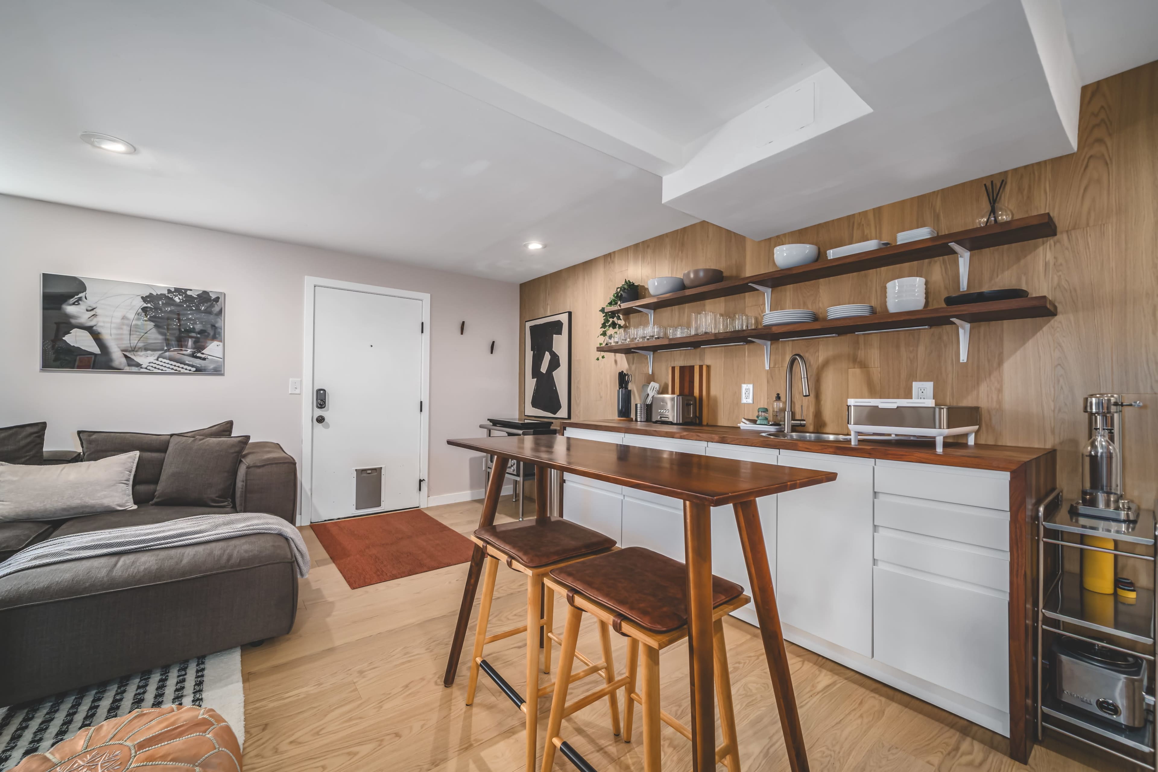 The image shows a compact apartment interior featuring a living area with a sofa, a kitchen space with open shelves, and a small dining table with two stools.