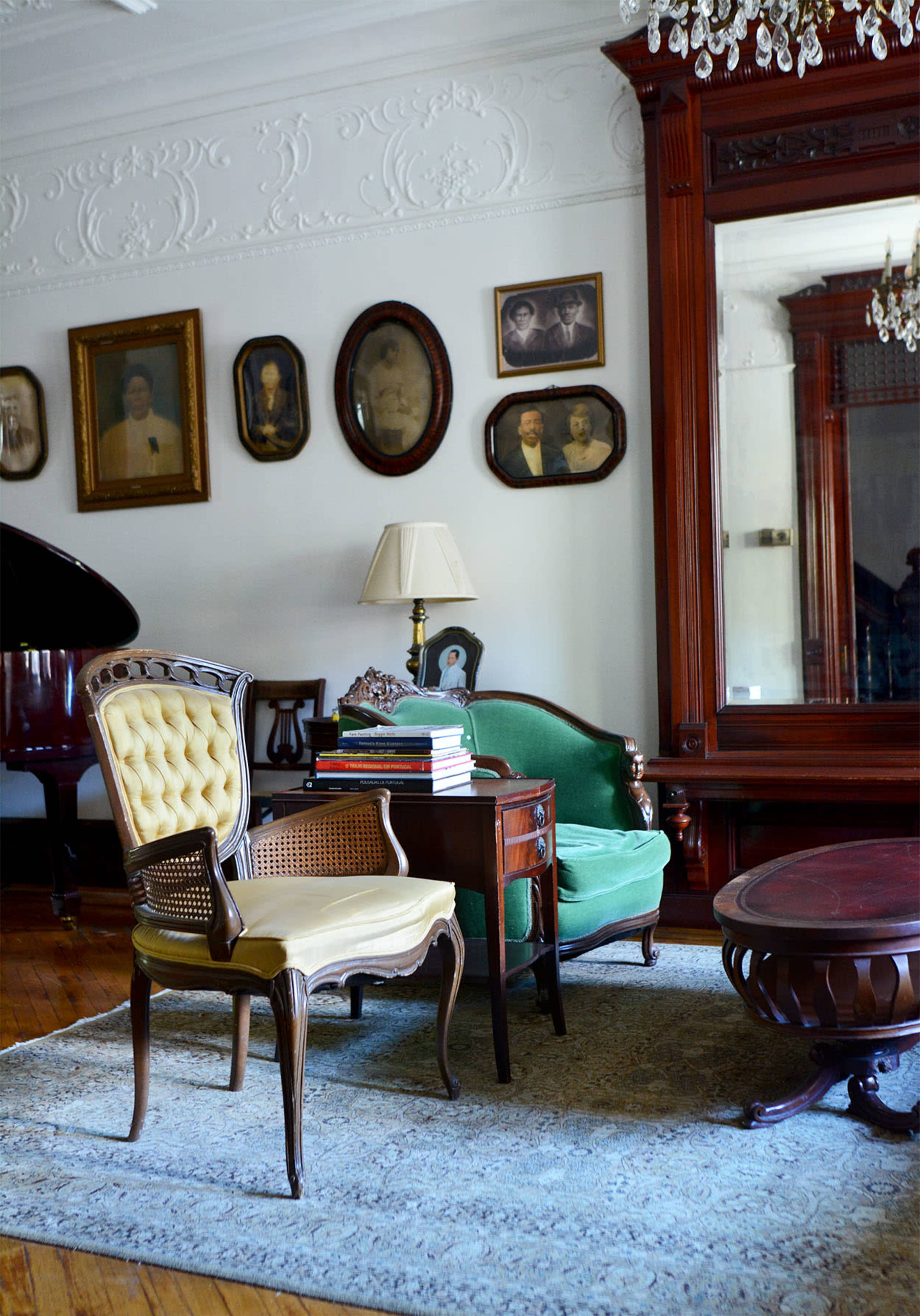The scene features an elegantly furnished room with a vintage green sofa, a yellow upholstered chair, a wooden table stacked with books, and framed portraits on the walls.
