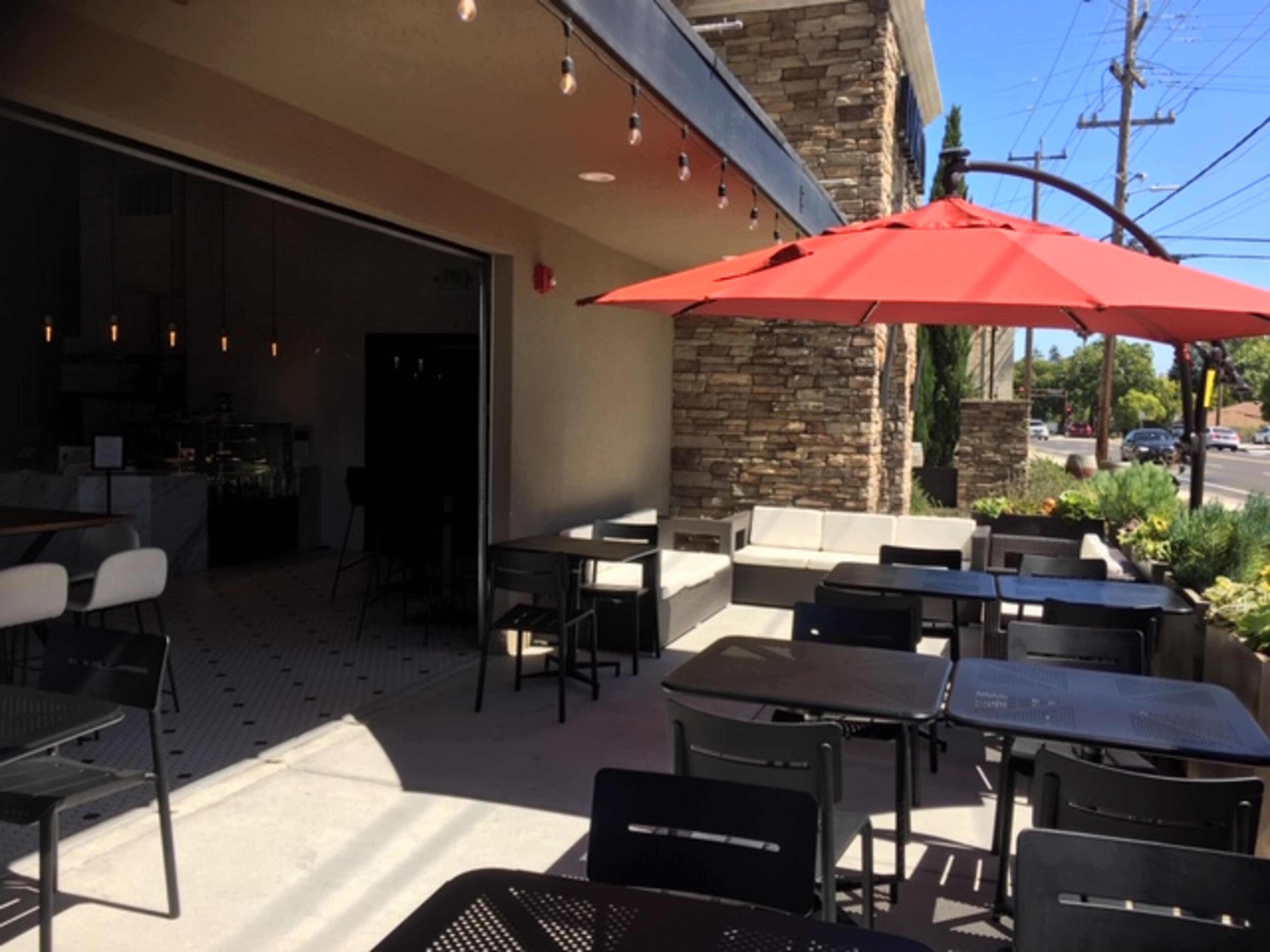 An outdoor seating area of a cafe with black tables and chairs, a red umbrella, and a stone wall in the background.