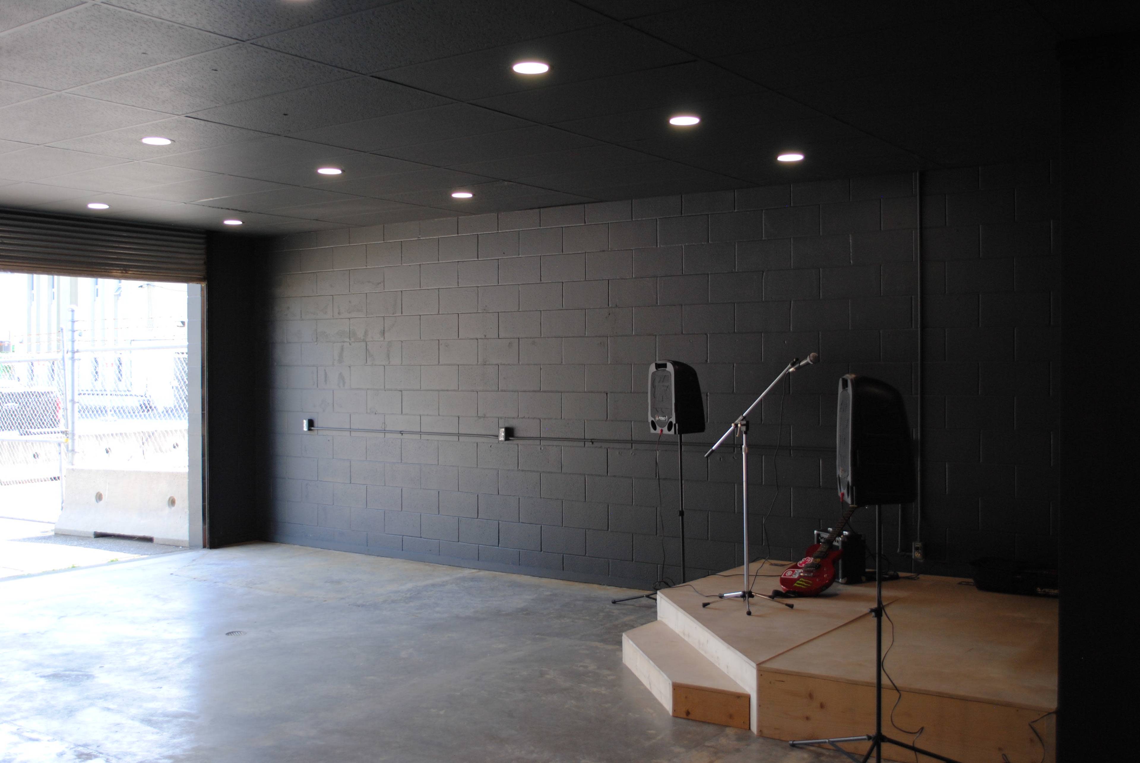 The image shows an empty room with a wooden stage, a microphone stand, and a guitar positioned on the floor, illuminated by overhead lights.
