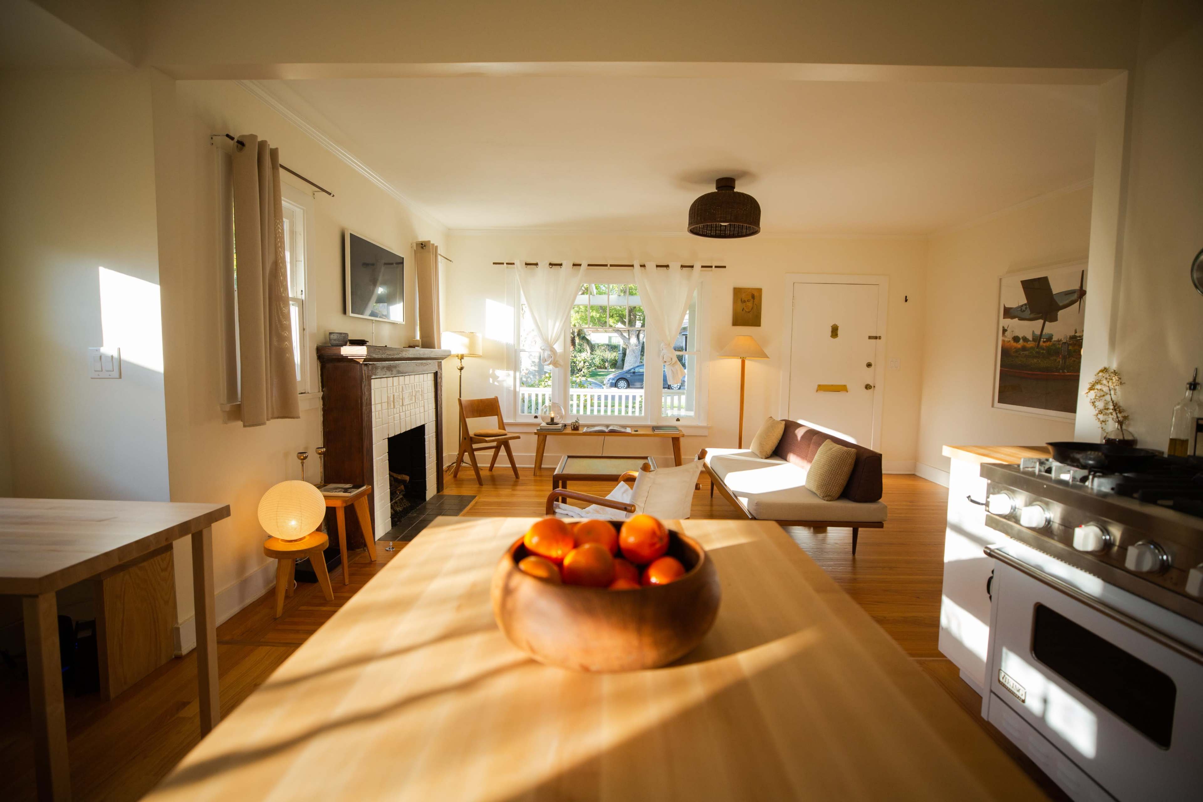 A bright, open living space features a wooden dining table with a bowl of oranges at its center, a fireplace, and a cozy seating area.