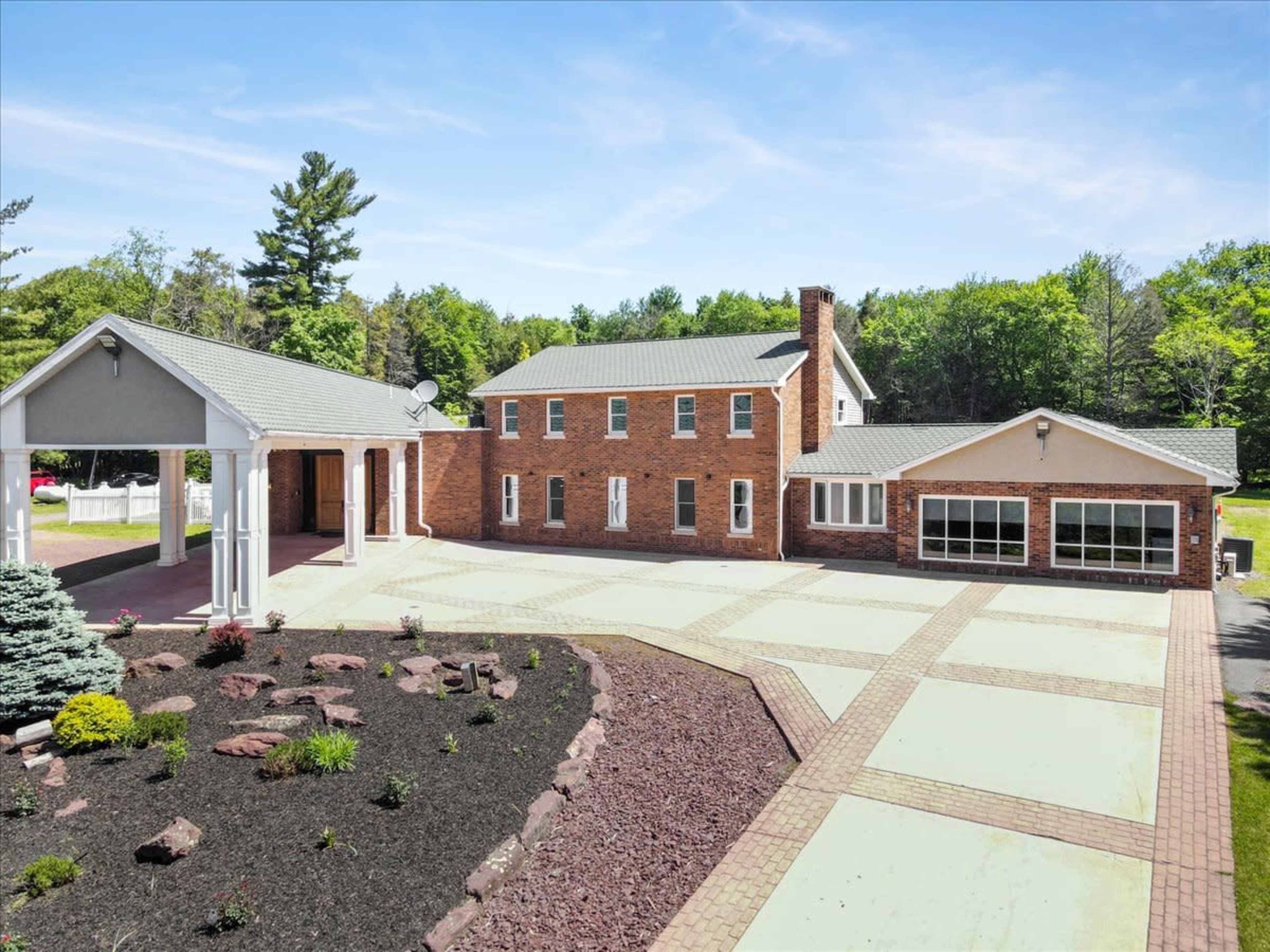 A two-story brick house with a covered porch, a large driveway, and landscaped gardens in a wooded setting.