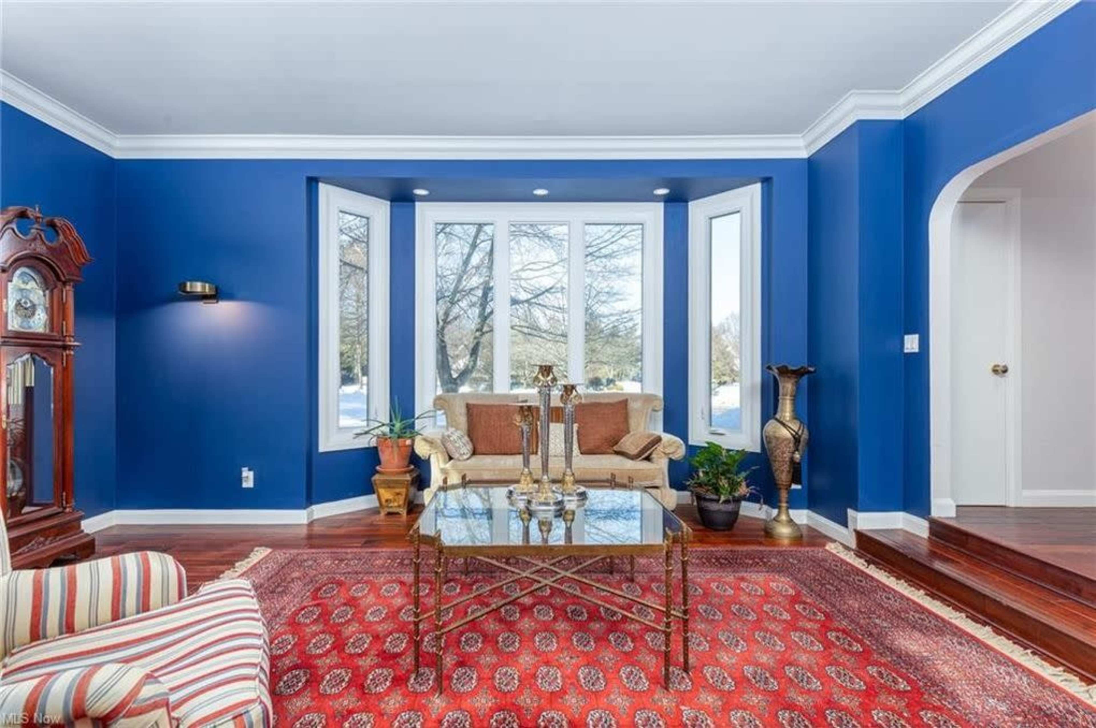 A living room with blue walls, a glass coffee table in the center, and a seating area featuring a sofa and armchair beside bay windows.