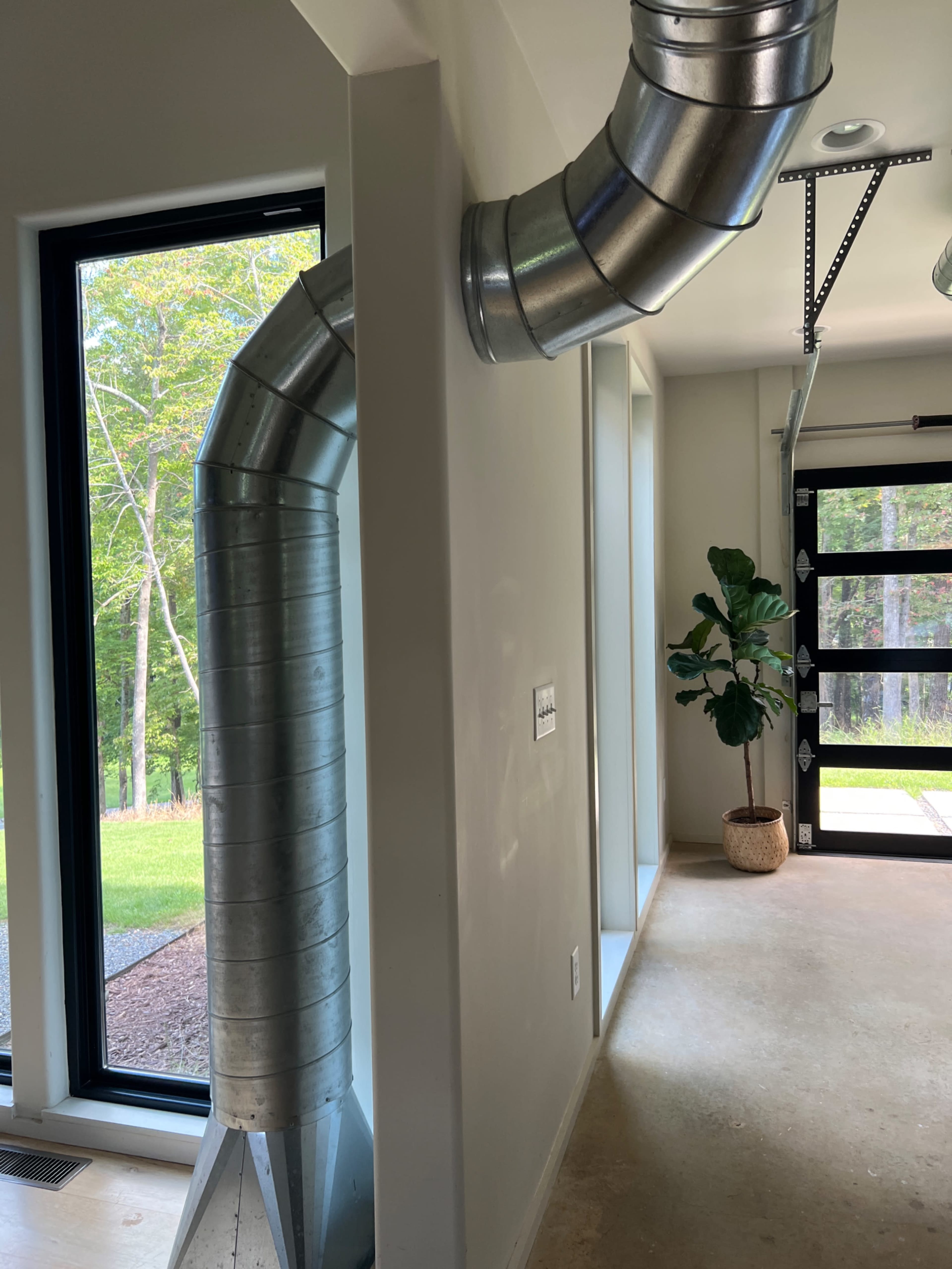 A metallic duct extends from the wall, curving toward a window with a view of greenery, and a garage door is visible on the right side of the image.