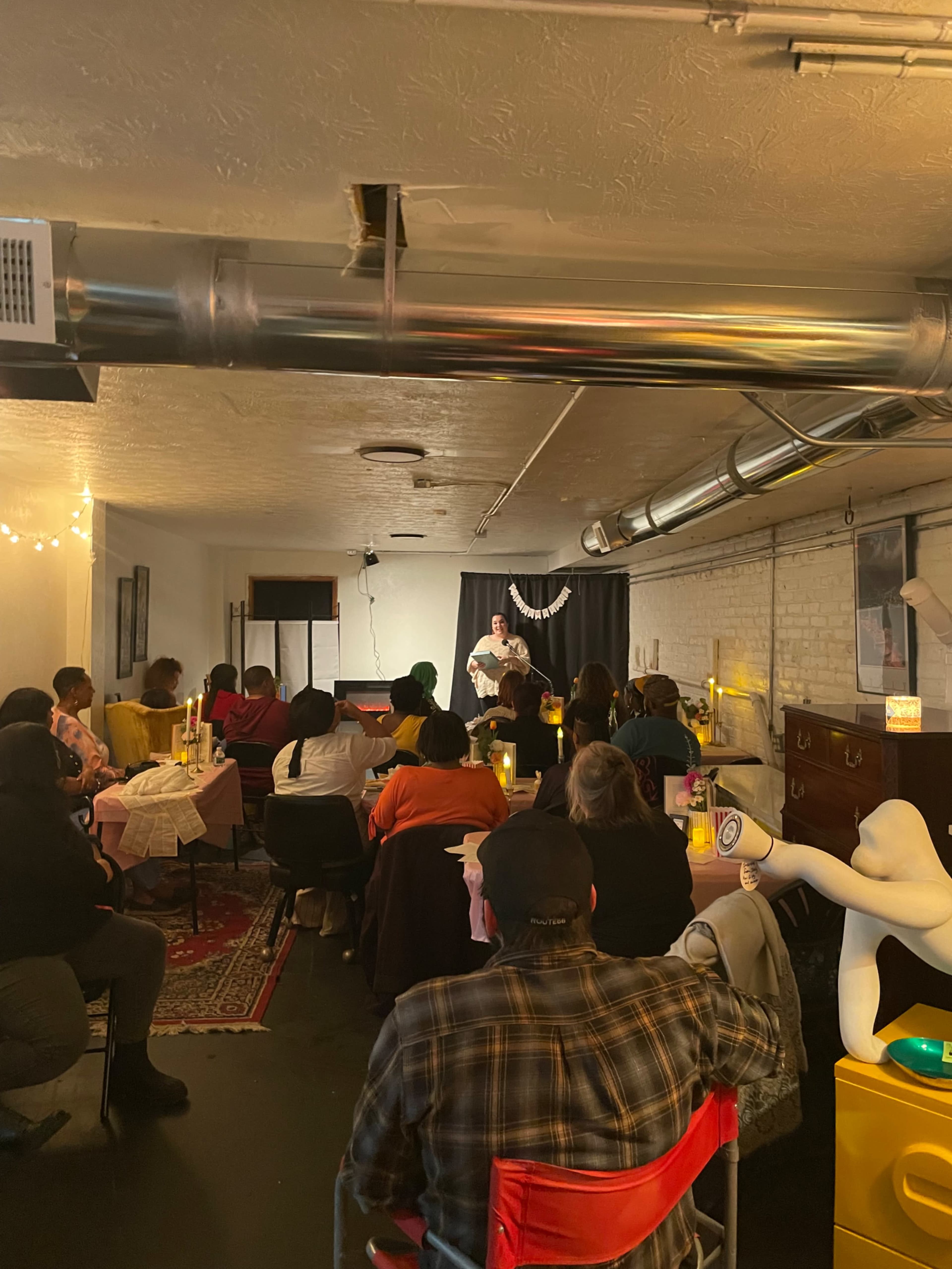 A comedian performs on stage in a dimly lit room filled with seated audience members at tables.
