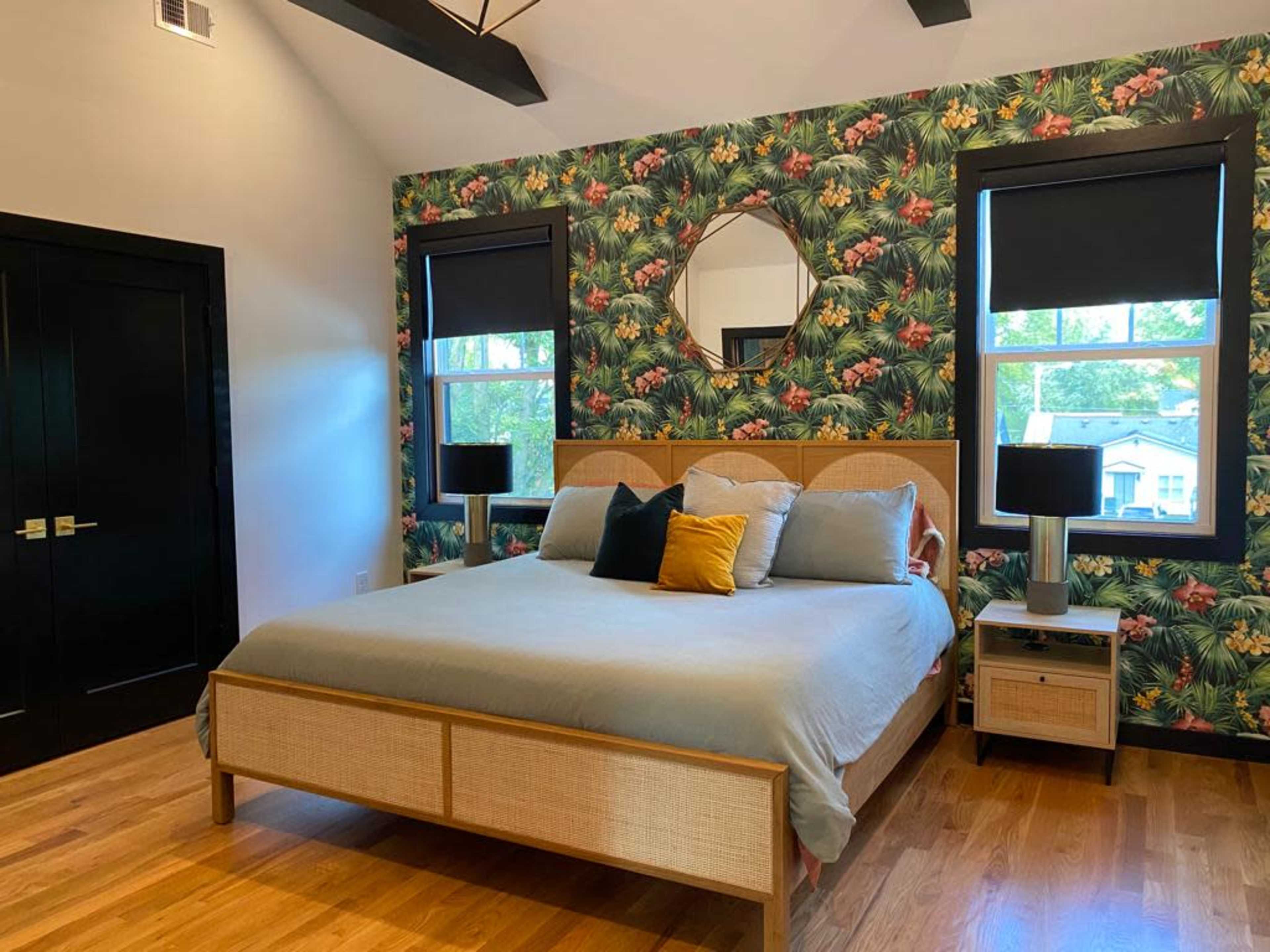 A bedroom featuring a bed with a light blue comforter, surrounded by tropical-themed wallpaper, two black lamps, and a large round mirror.