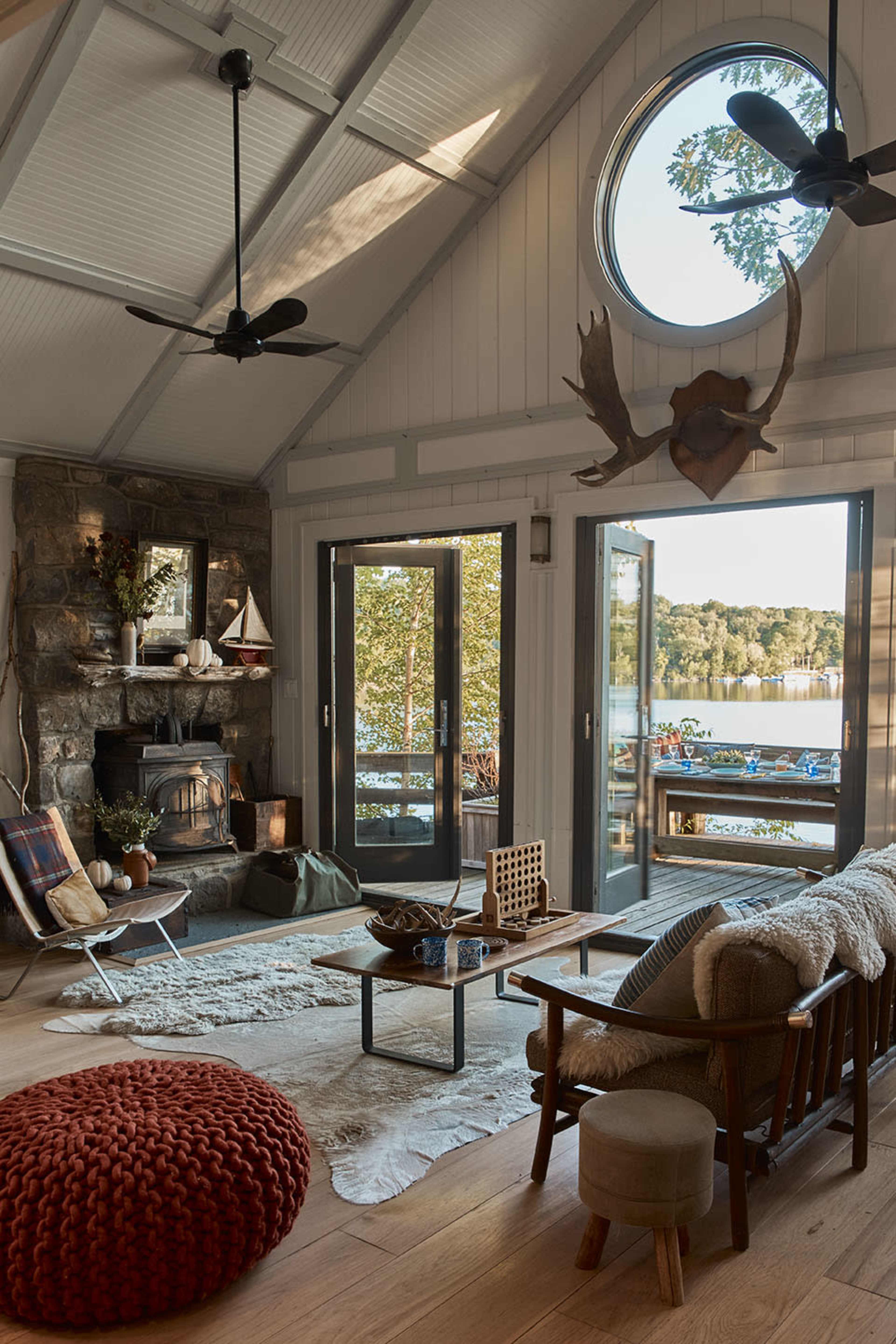 The living room features large windows overlooking a lake, a stone fireplace, and a mix of rustic and modern furniture.