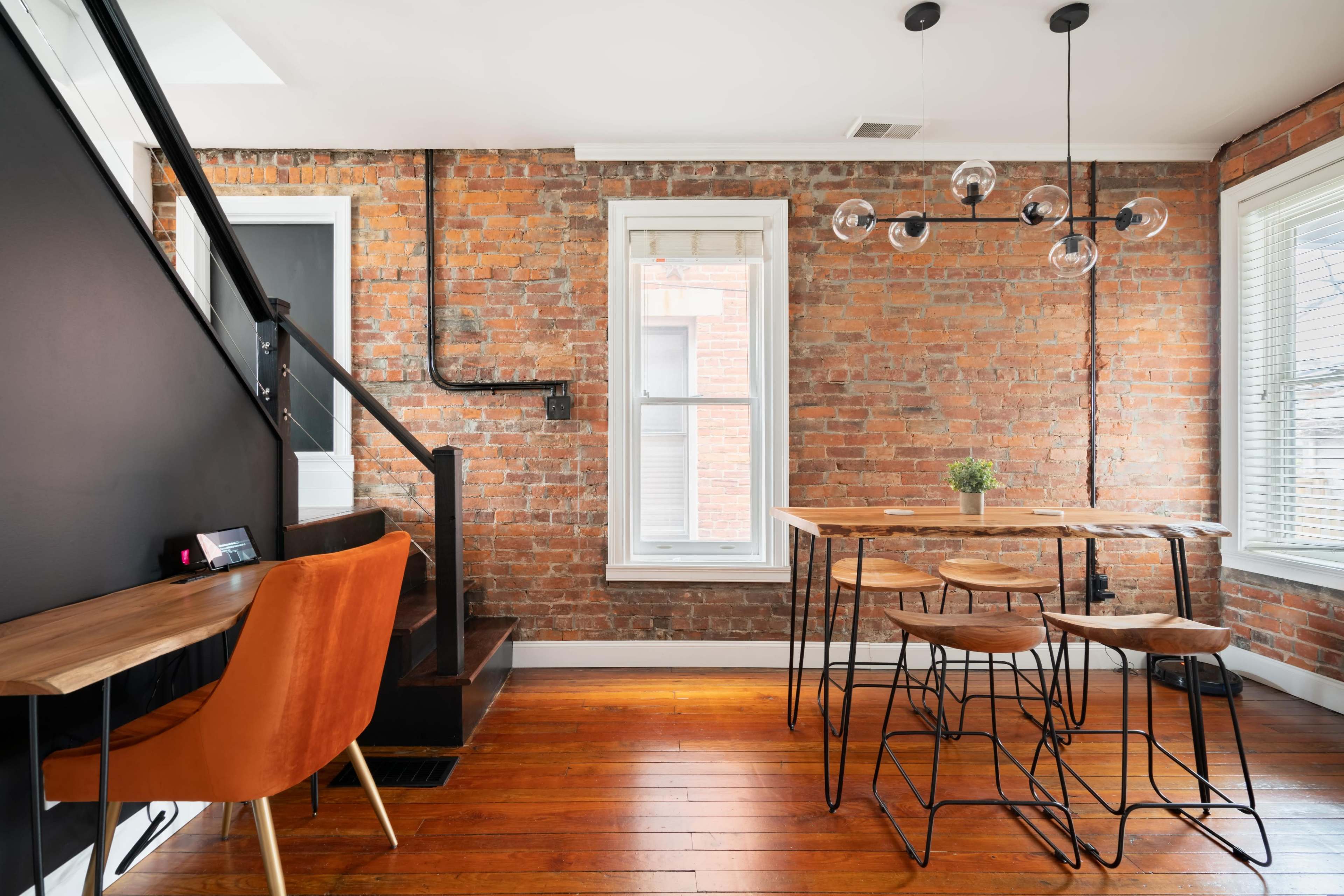 A modern dining area with a wooden table surrounded by metal chairs, set against a backdrop of exposed red brick walls and large windows.