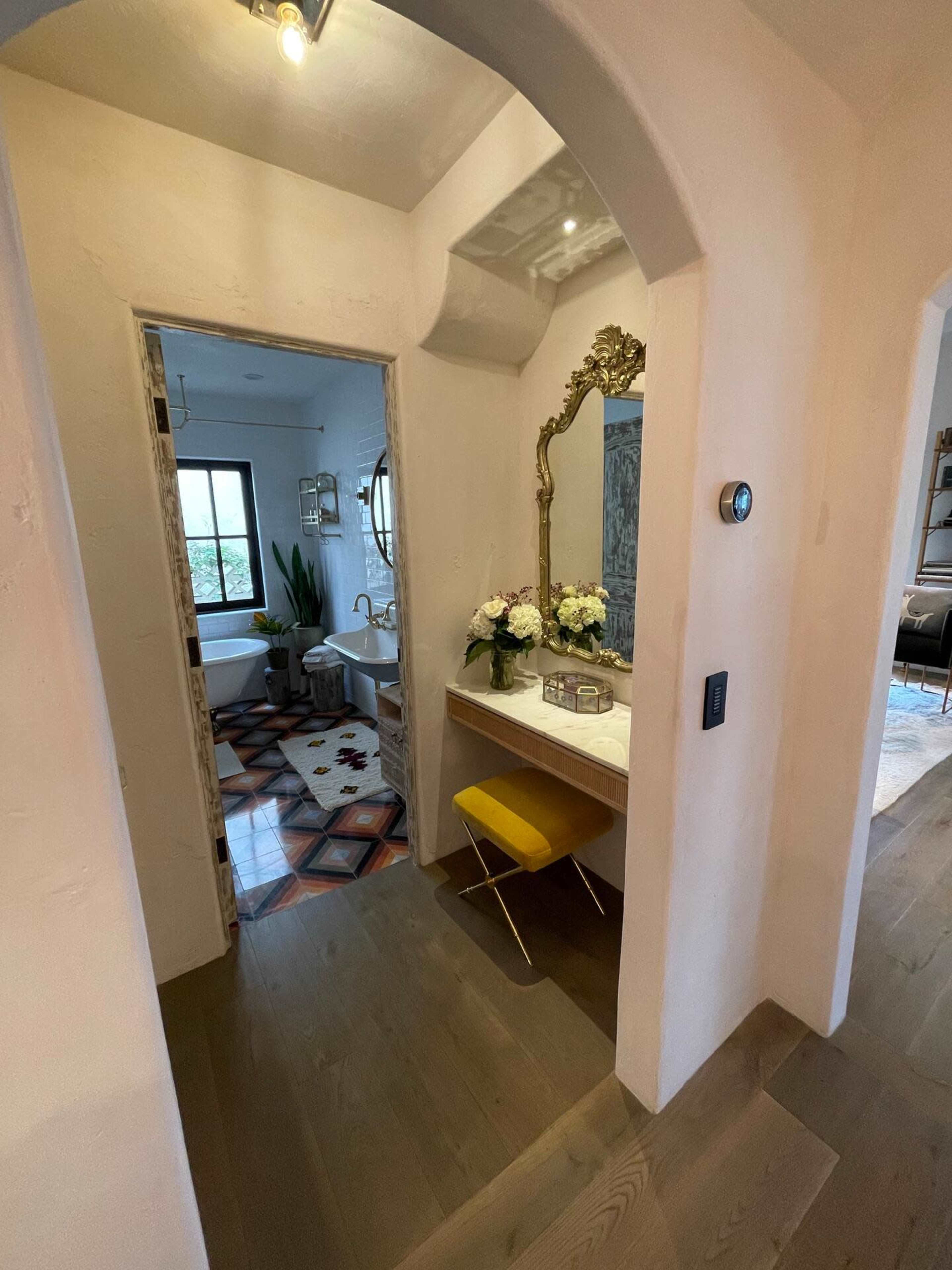 A hallway leading to a bathroom, featuring a decorative mirror, a small vanity with a vase of flowers, and a stylish yellow chair.