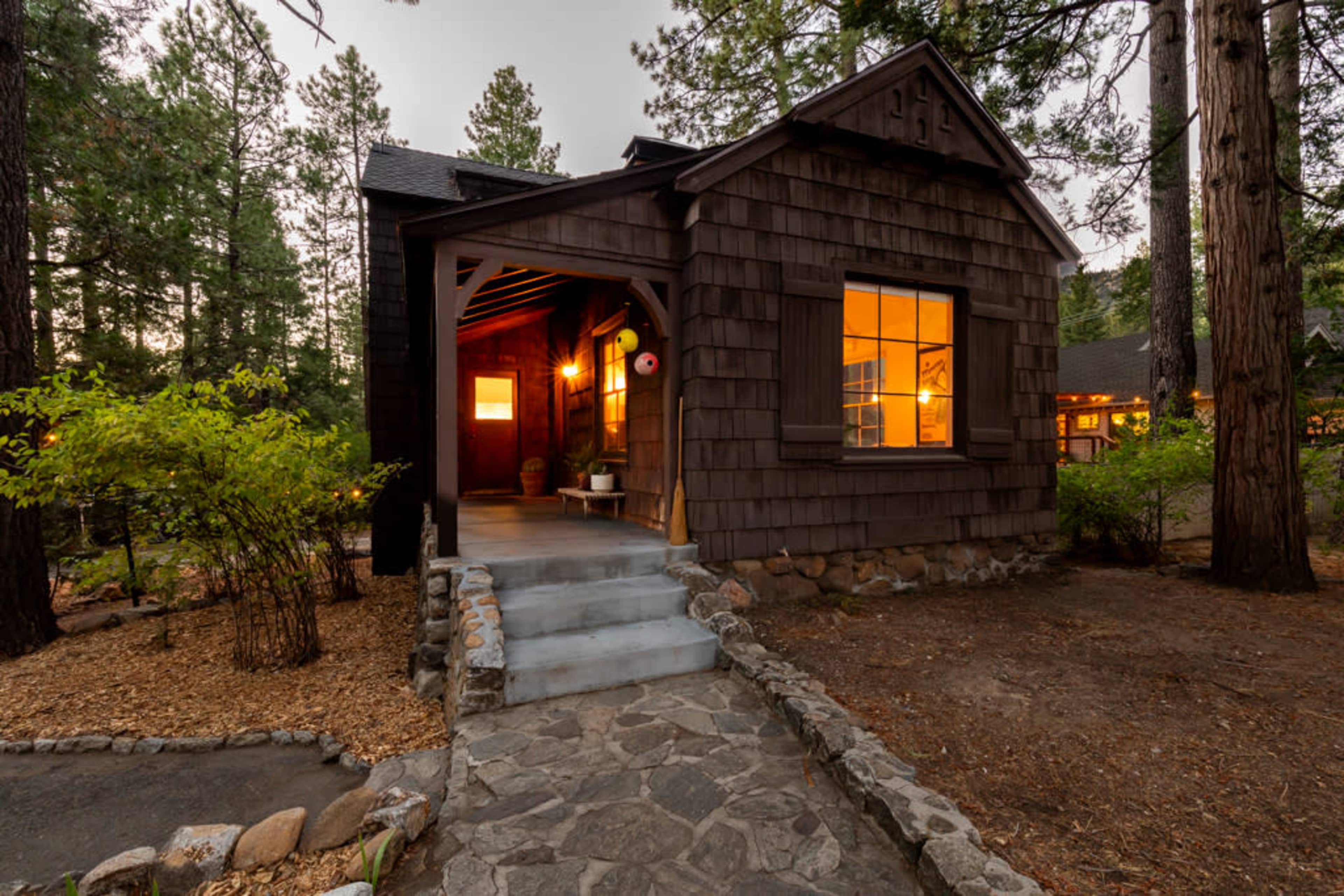A small, rustic cabin with dark wood siding, a stone walkway, and warm lights glowing from the windows is surrounded by pine trees.