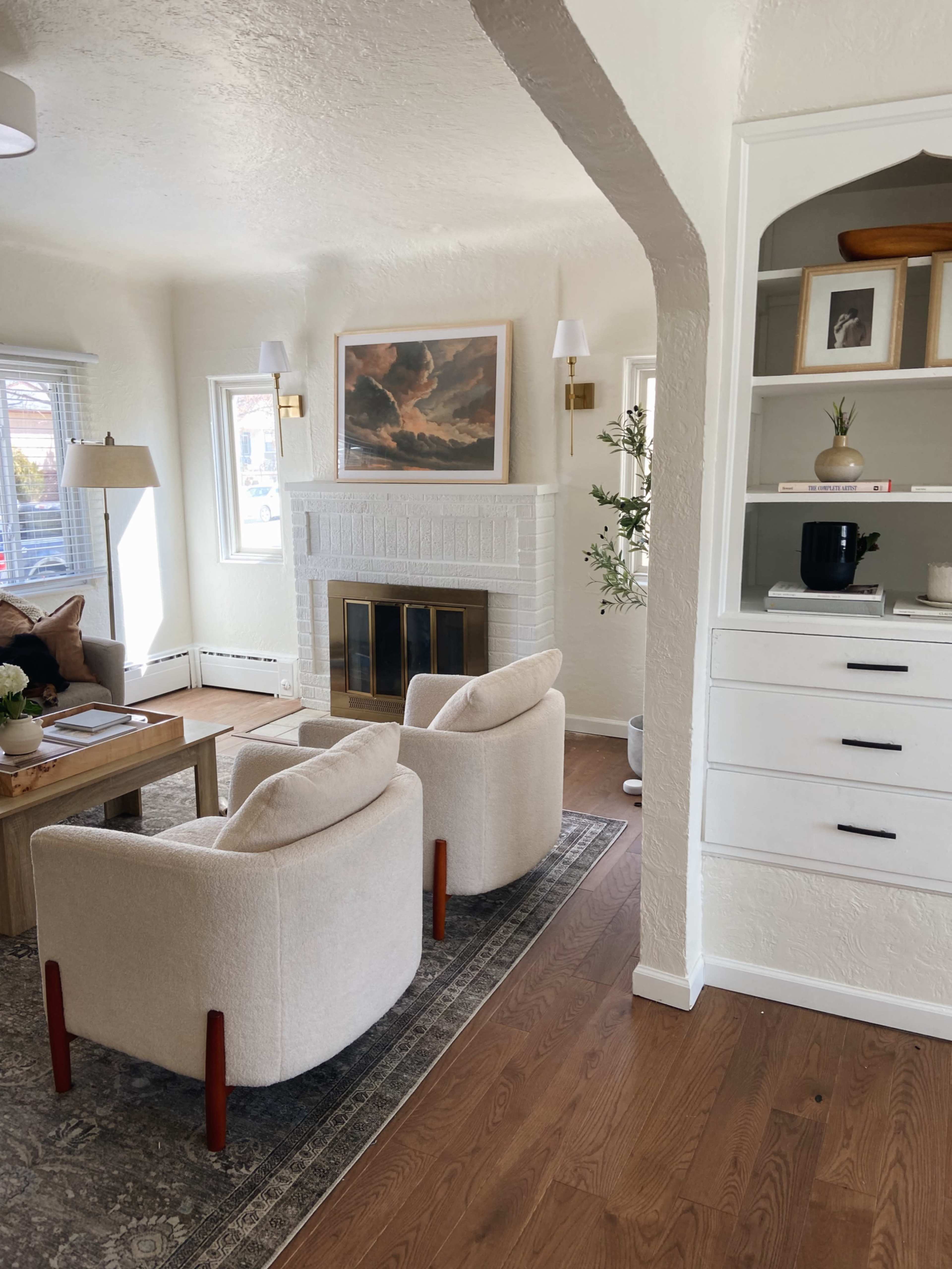 The image shows a cozy living room featuring two beige armchairs, a wooden coffee table, a fireplace, and a shelf with decorative items.