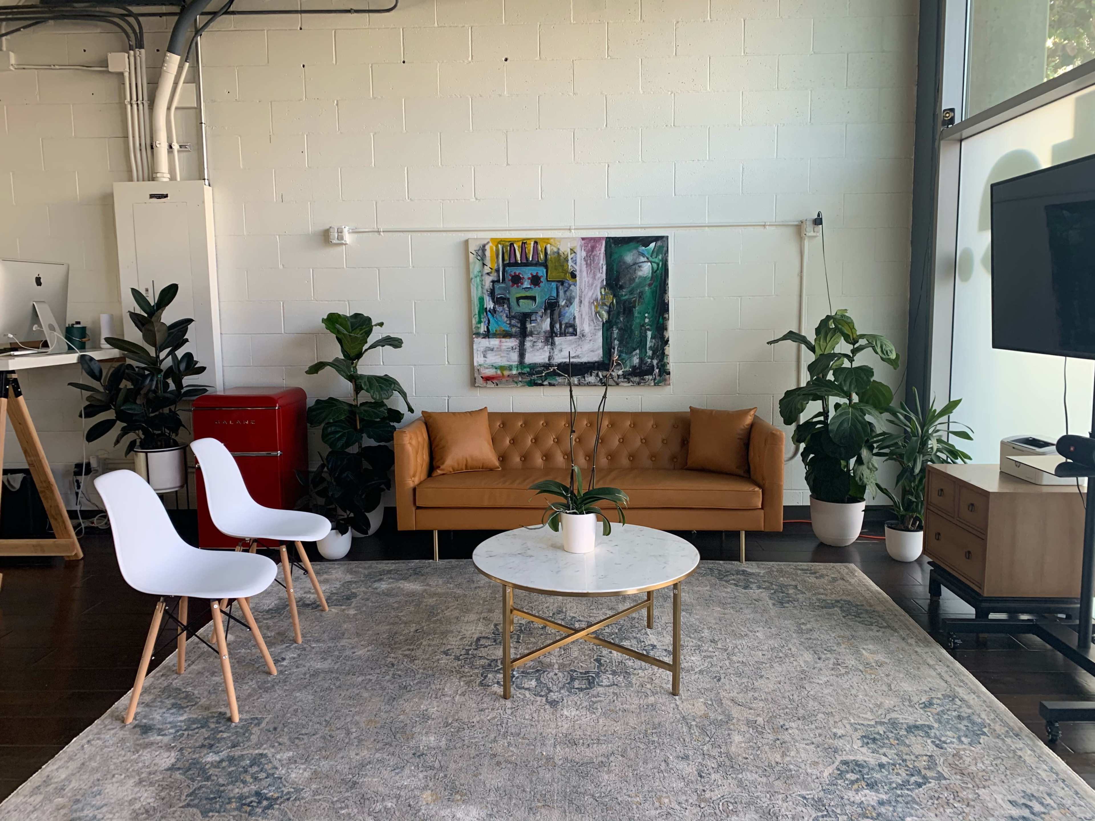 A modern living space featuring a brown sofa, a round marble coffee table, two white chairs, and various plants, all set against a light-colored wall with a colorful artwork.