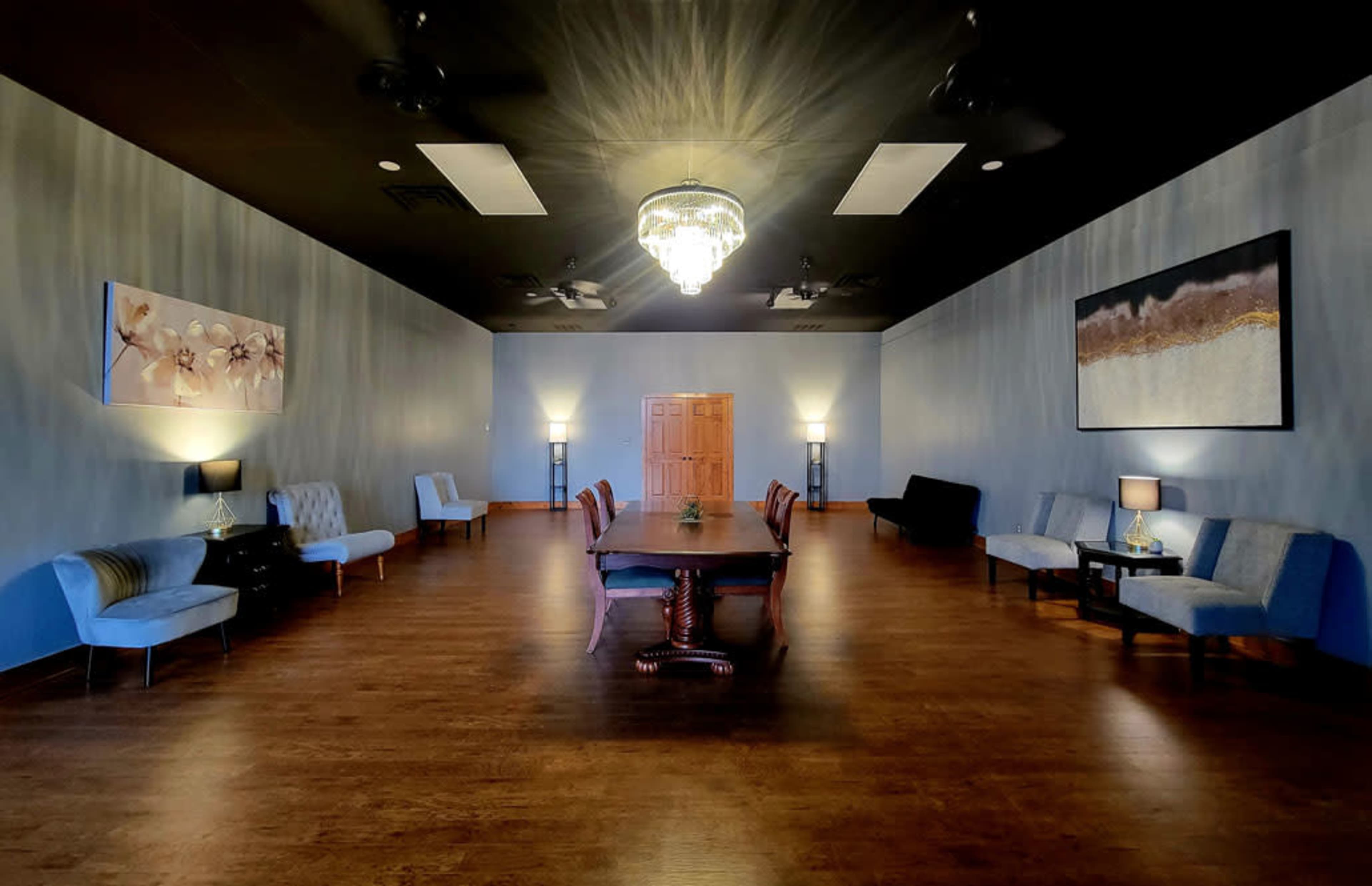 The image shows a spacious, well-lit room with a long wooden table in the center, surrounded by various seating arrangements and large artworks on the walls.