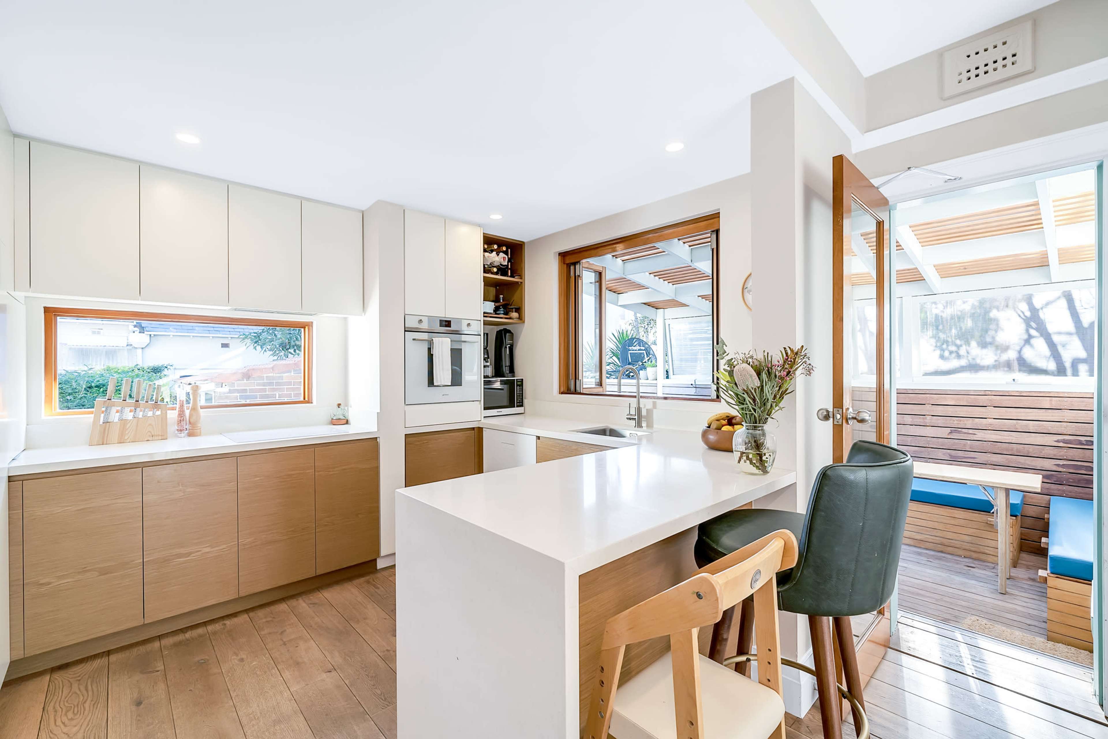 A modern kitchen with wooden accents, a white countertop, and large windows providing natural light.