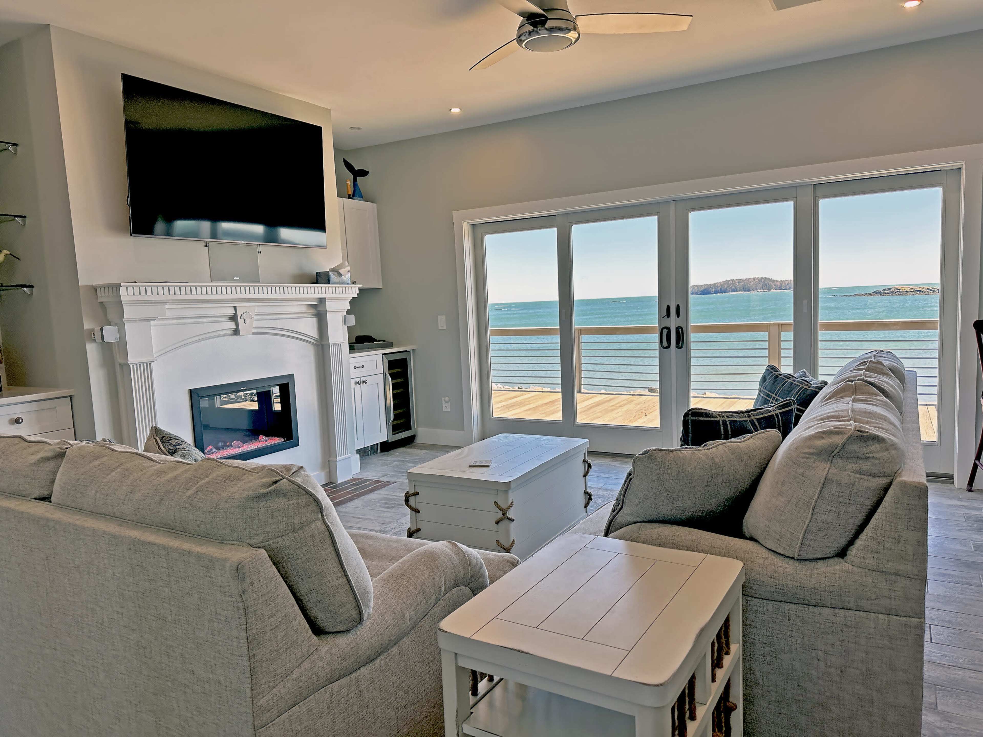 The image shows a light-filled living room with a gray sectional sofa, a modern fireplace, and large sliding glass doors that open to a view of the water and a distant island.