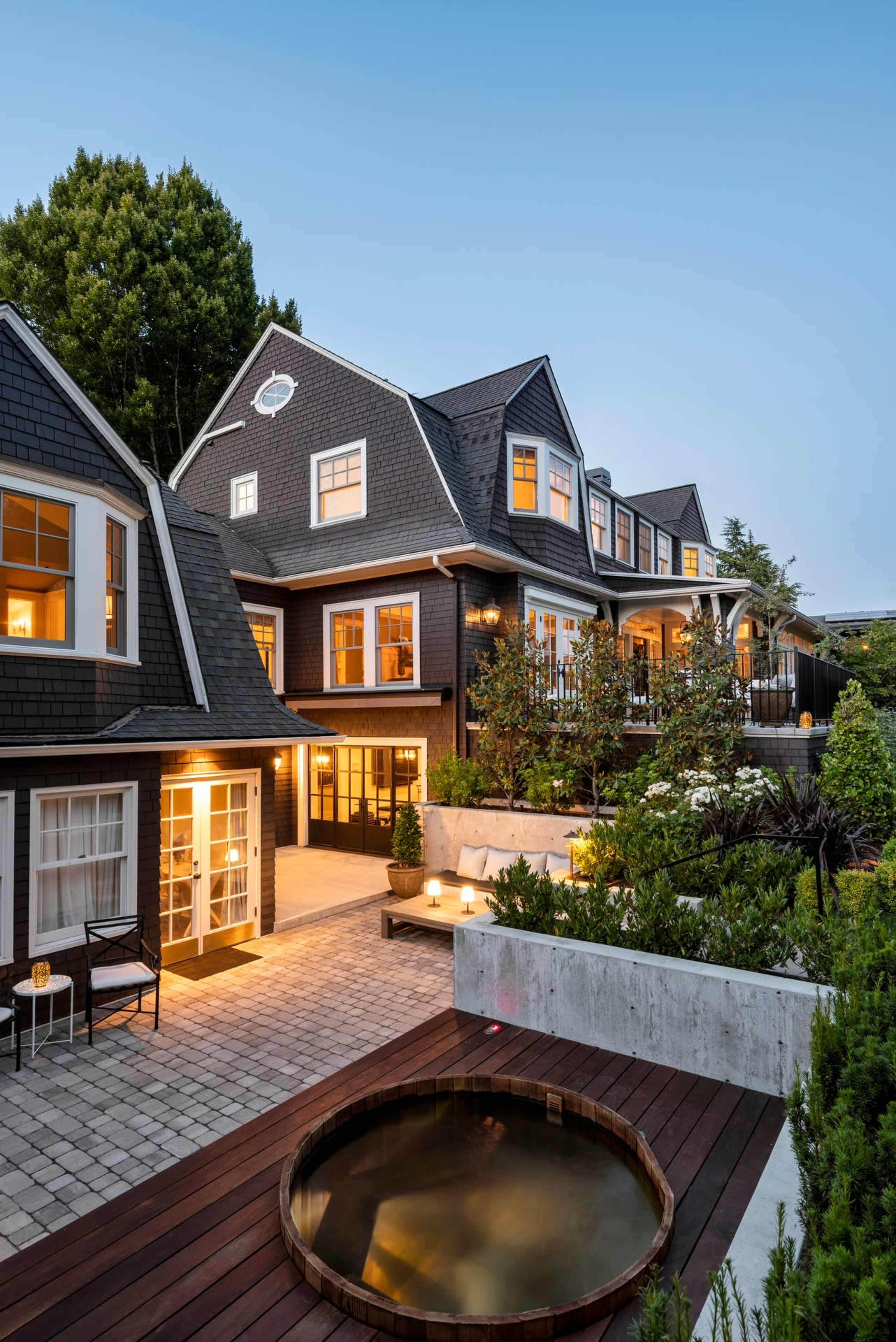 A modern, multi-story house with dark shingles features a patio and a wooden hot tub surrounded by landscaped greenery and soft outdoor lighting.